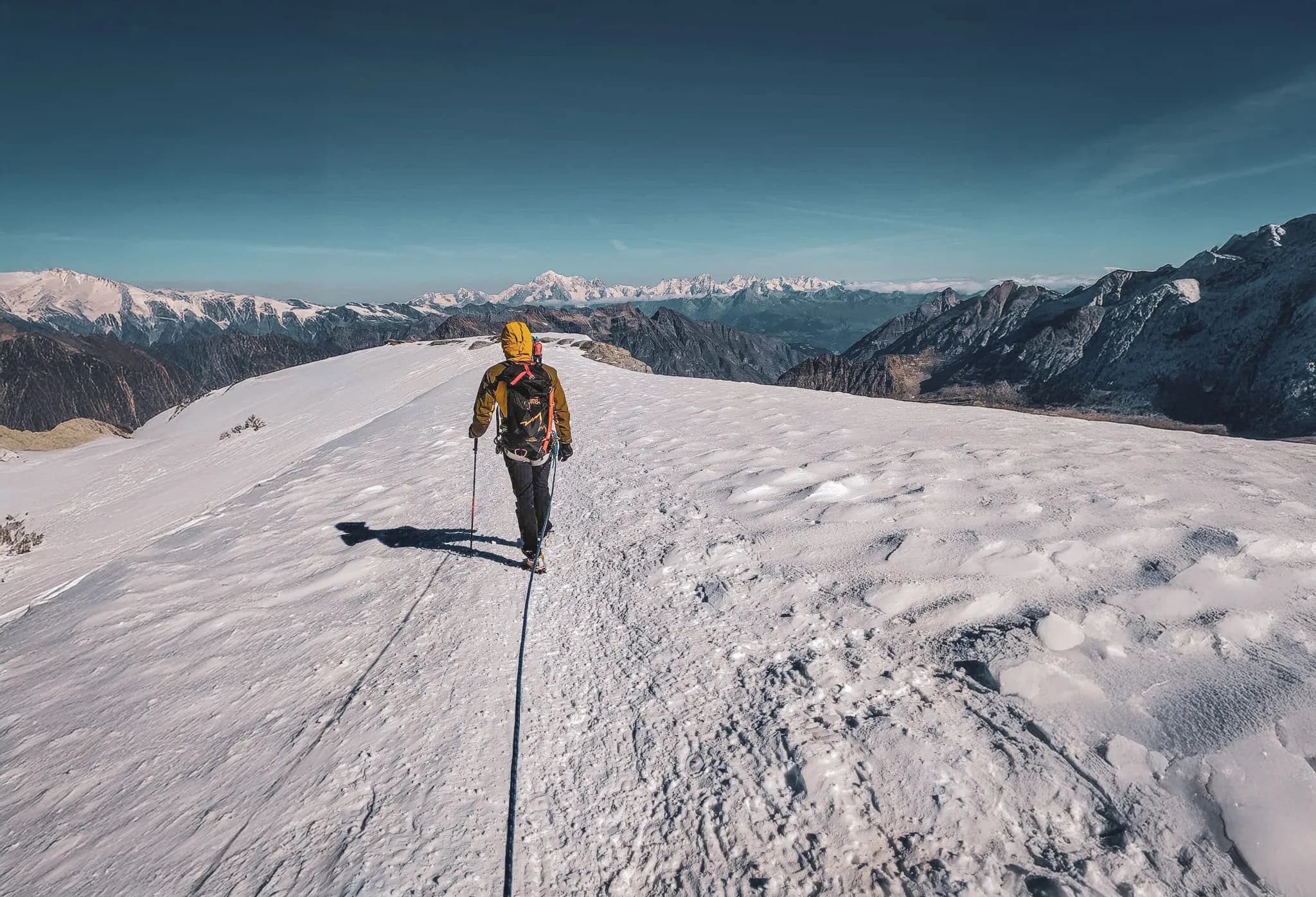 A mountaineer walks on a glacier, surrounded by majestic mountains under a blue sky.