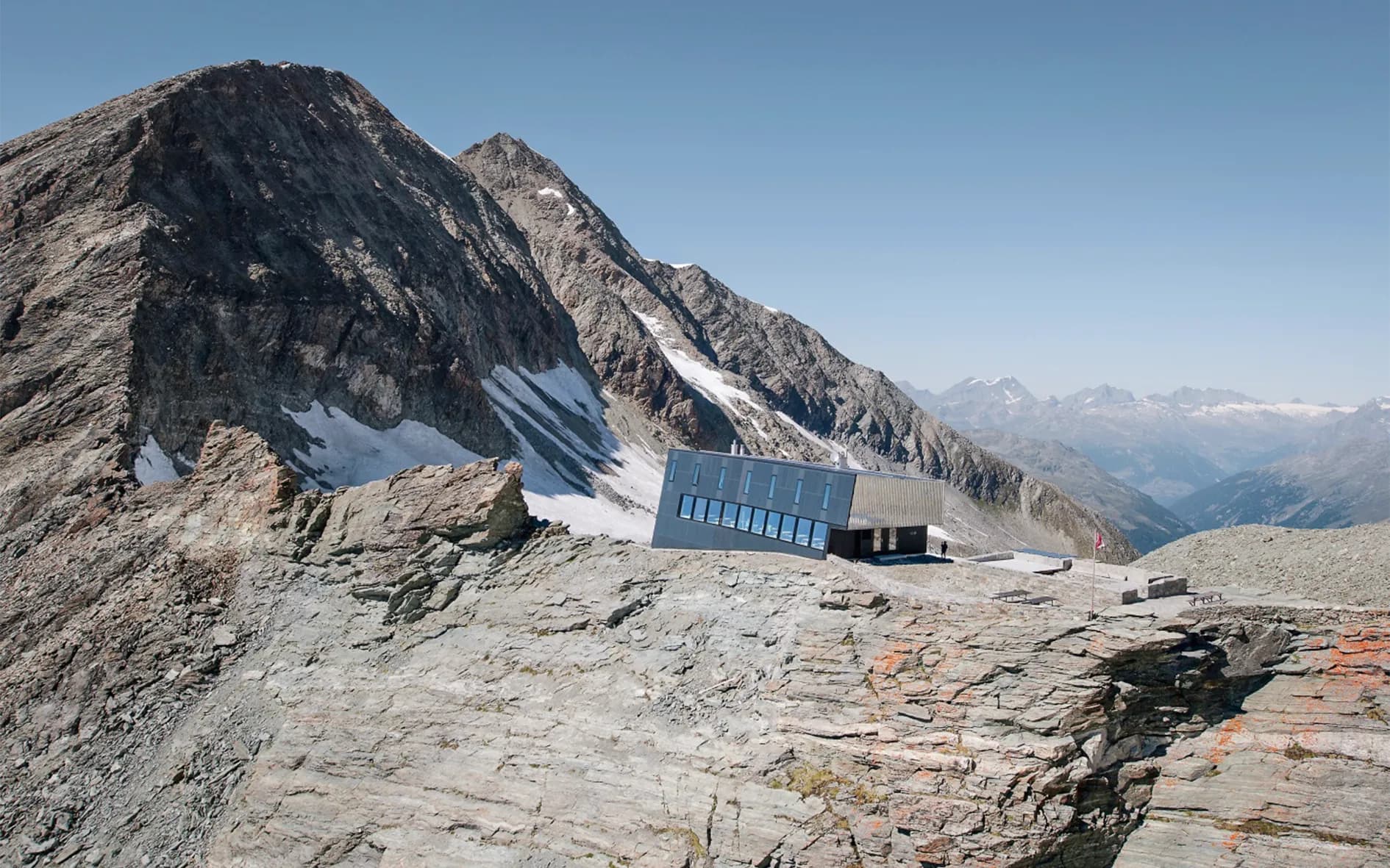 A modern, angular building sits on a mountain ridge, surrounded by craggy rocky landscapes and majestic peaks.