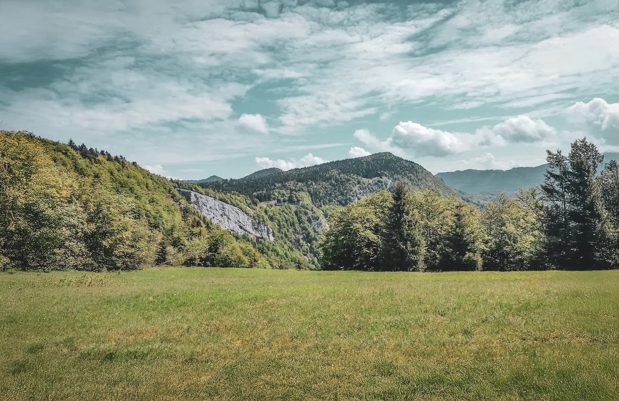 A mountainous landscape with steep ridges and green slopes. The sunlight illuminates the cliffs, highlighting the rock strata. In the background, a valley unfolds