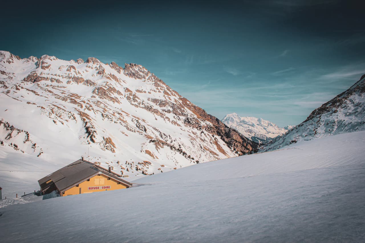 A mountain refuge is in the foreground, surrounded by a snow-covered Alpine landscape. The surrounding area is dominated by majestic peaks, partially covered in snow, which rise up against a sky of a pale blue.