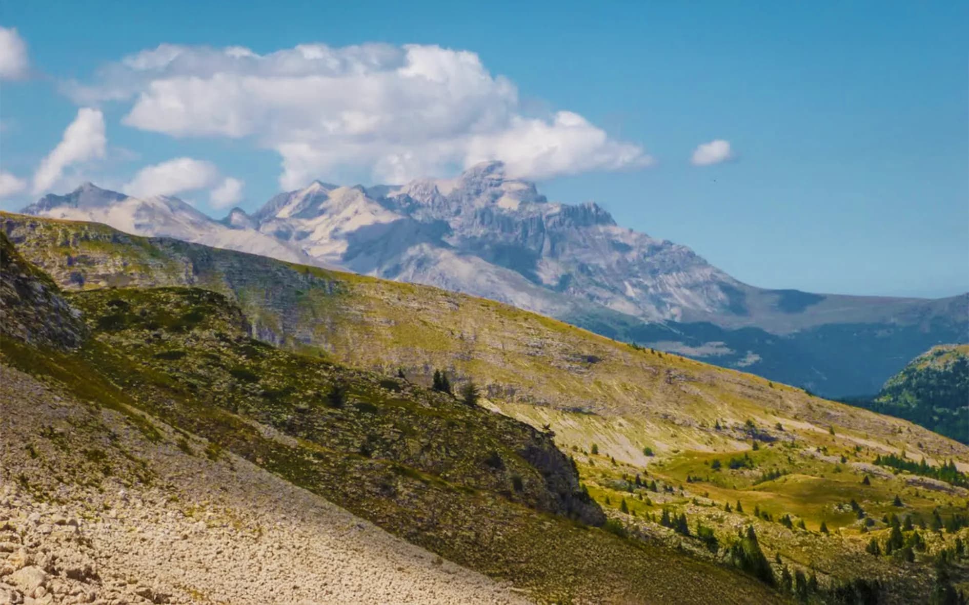 A mountainous landscape with green hills and rocky ridges. The majestic mountains rise up in the background, under a clear blue sky dotted with white clouds. The ground features textures