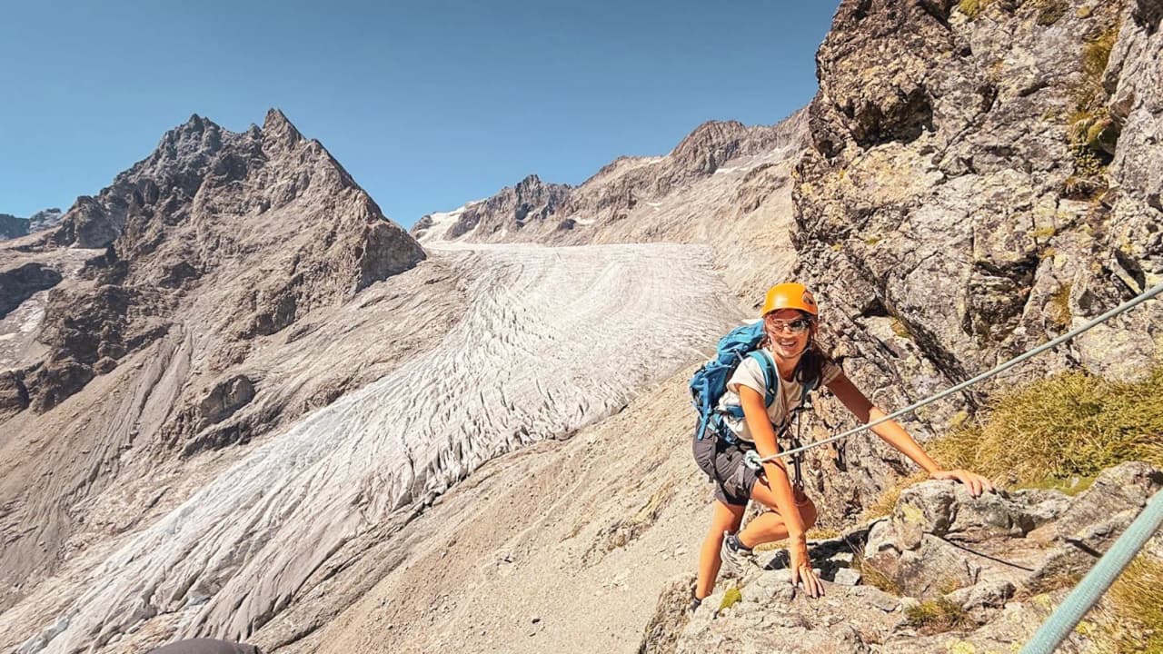 Grimpeuse souriante sur un sentier alpin, face à un magnifique glacier sous un ciel bleu.