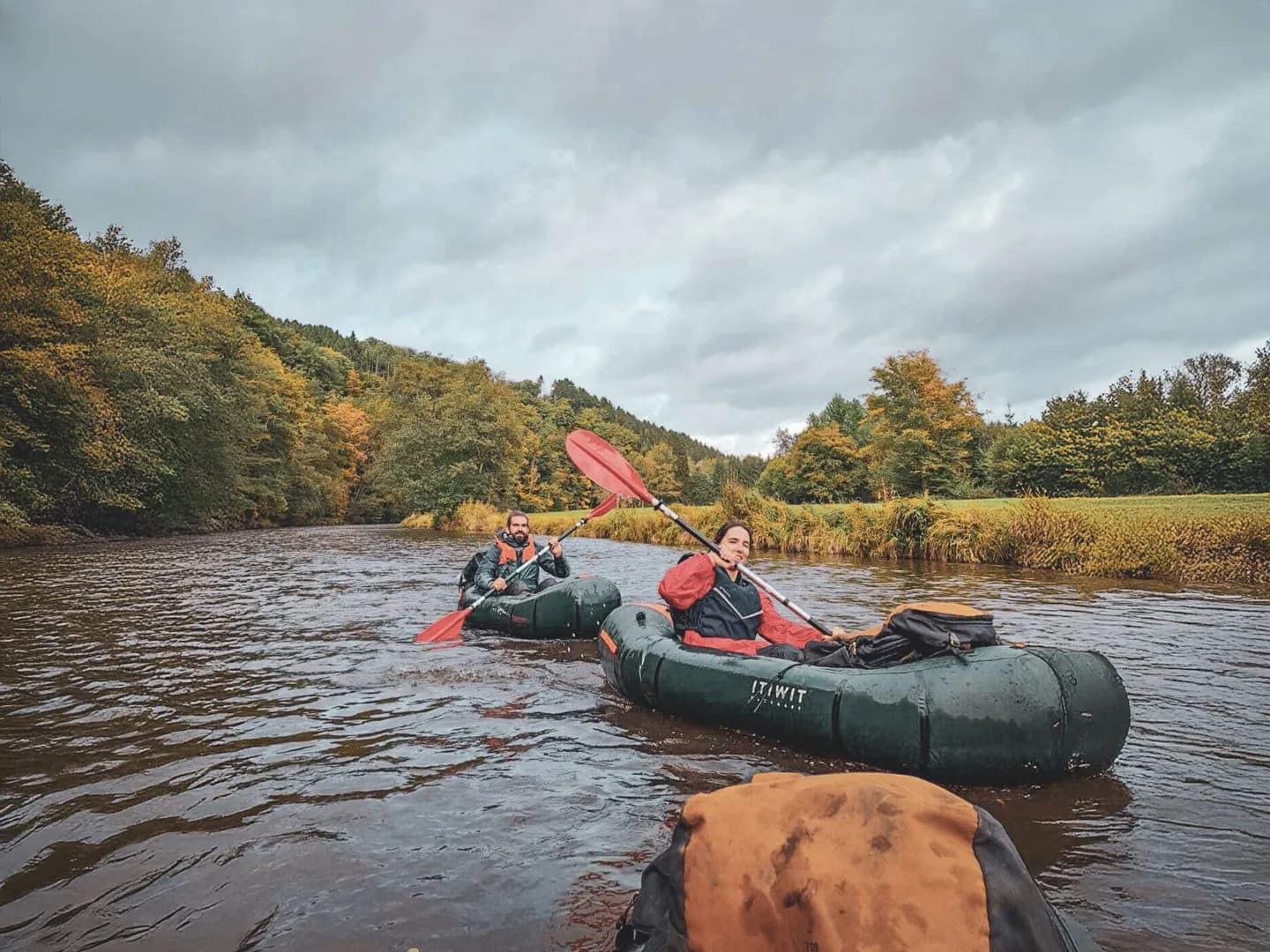 Two packrafters glide down a river surrounded by autumn-coloured forests.