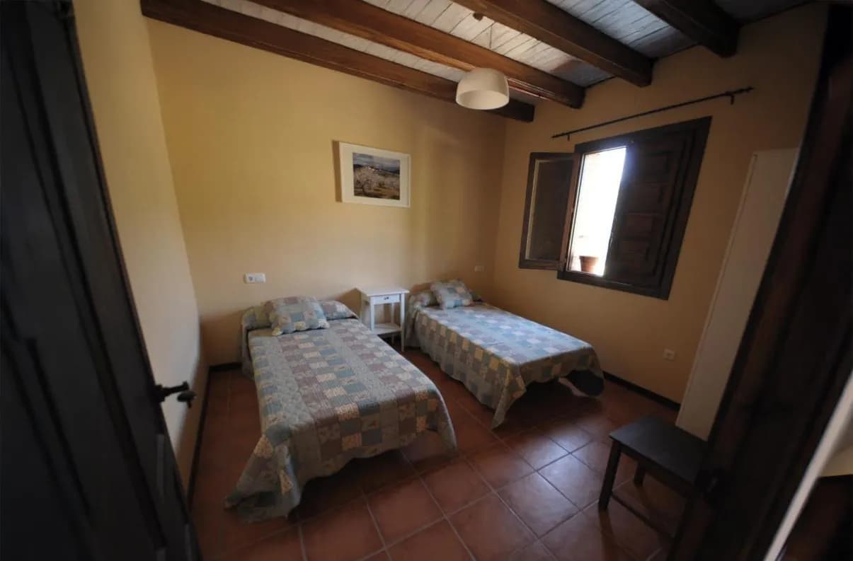 A cosy room with twin beds, each covered with a square-patterned blanket. The walls are painted beige, and the ceiling is accentuated by wooden beams. A picture on the wall adds