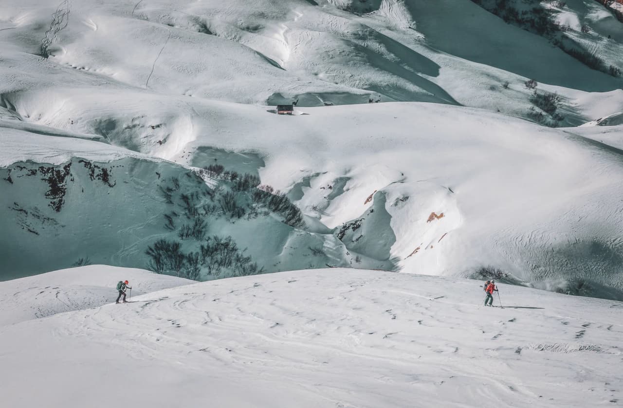 A snowy landscape with rolling hills covered in snow. Two skiers, one in a red jacket and the other in a dark jacket, are making their way down a slope. In the distance, a small cab