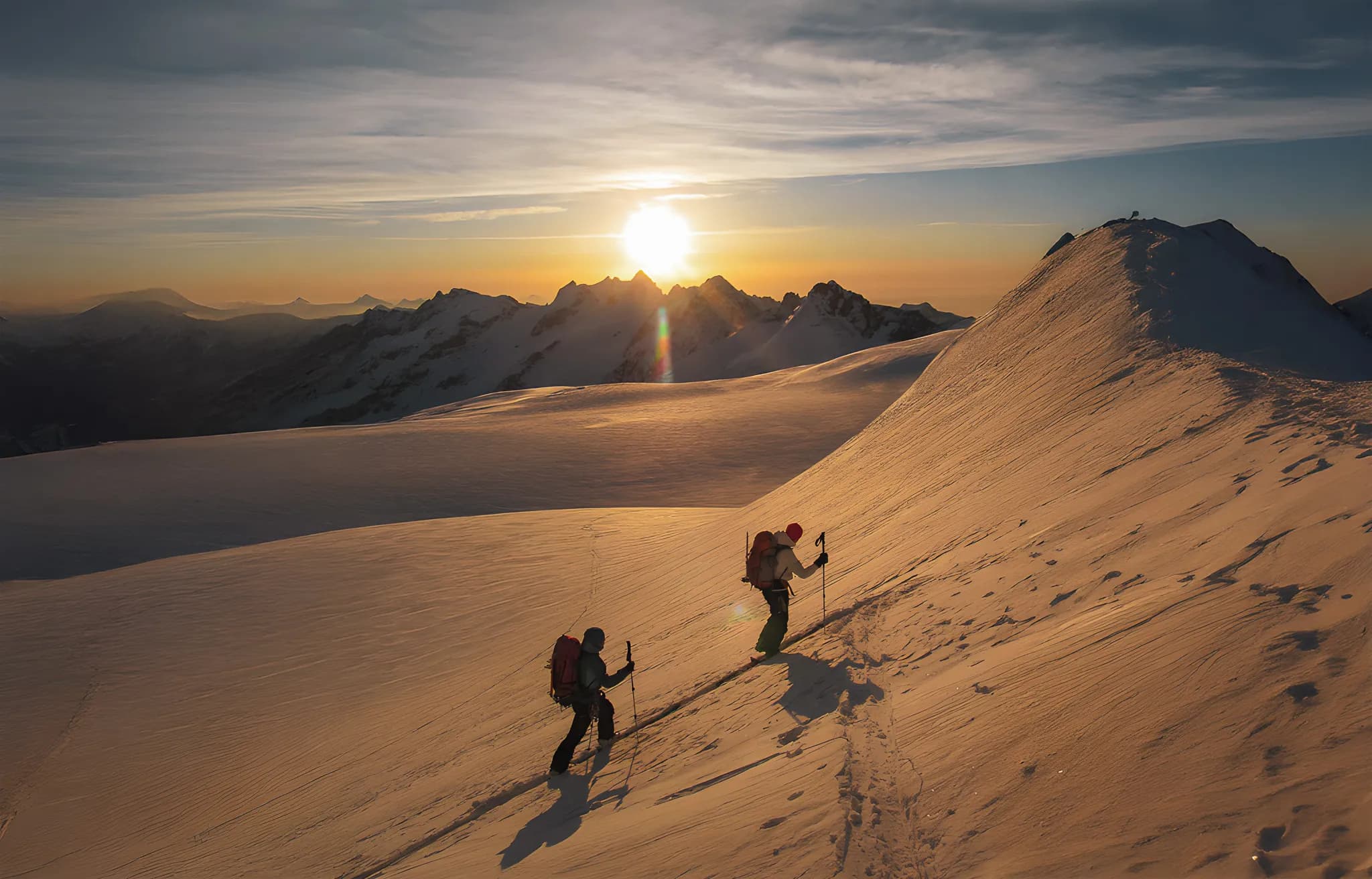 Deux skieurs ascensionnistes gravissent des pentes enneigées au lever du soleil, entourés de montagnes majestueuses.