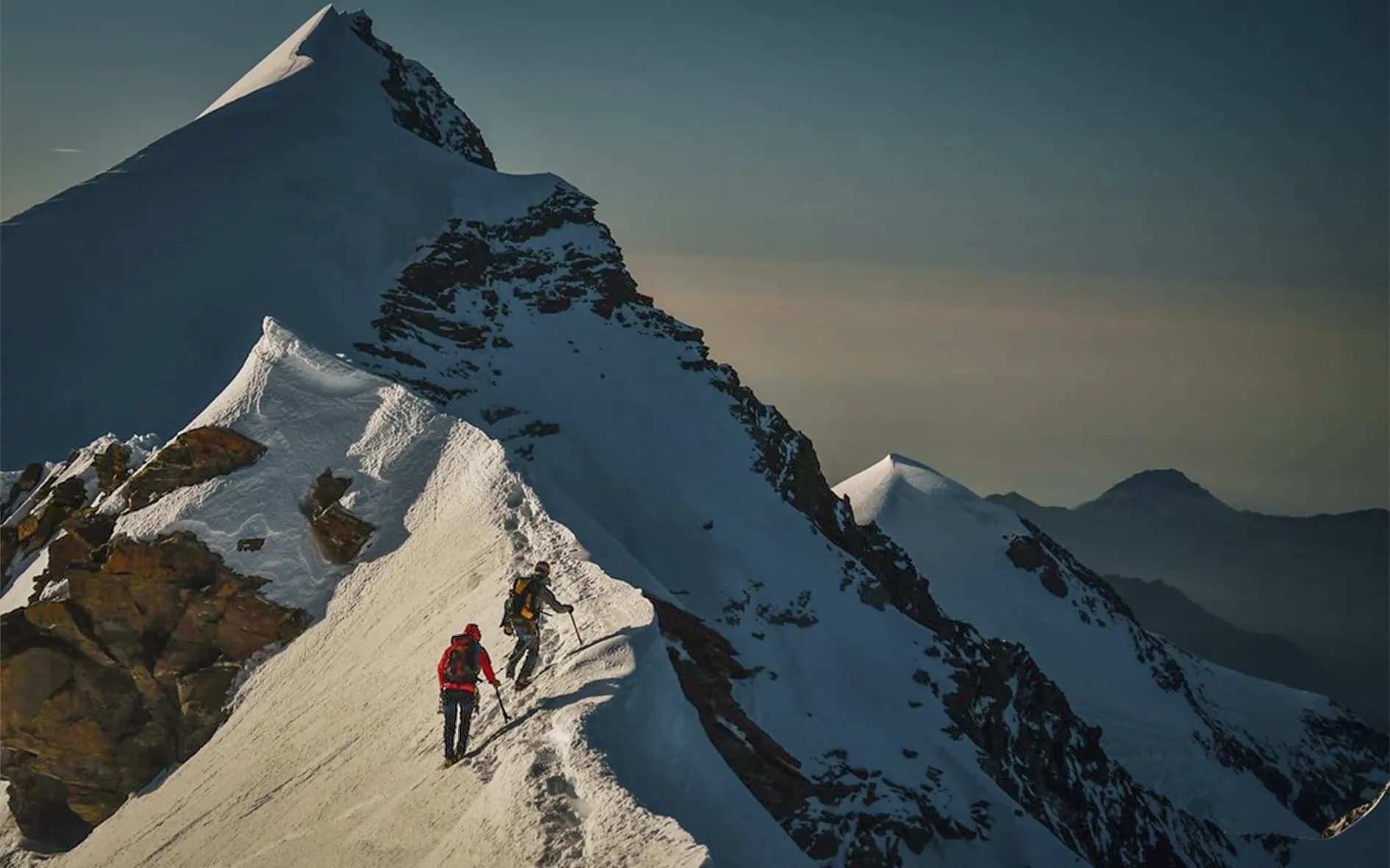 A group of climbers making their way up a snow-covered mountain peak. They are equipped with mountaineering gear, such as ice axes and ropes, and are wearing safety helmets. At