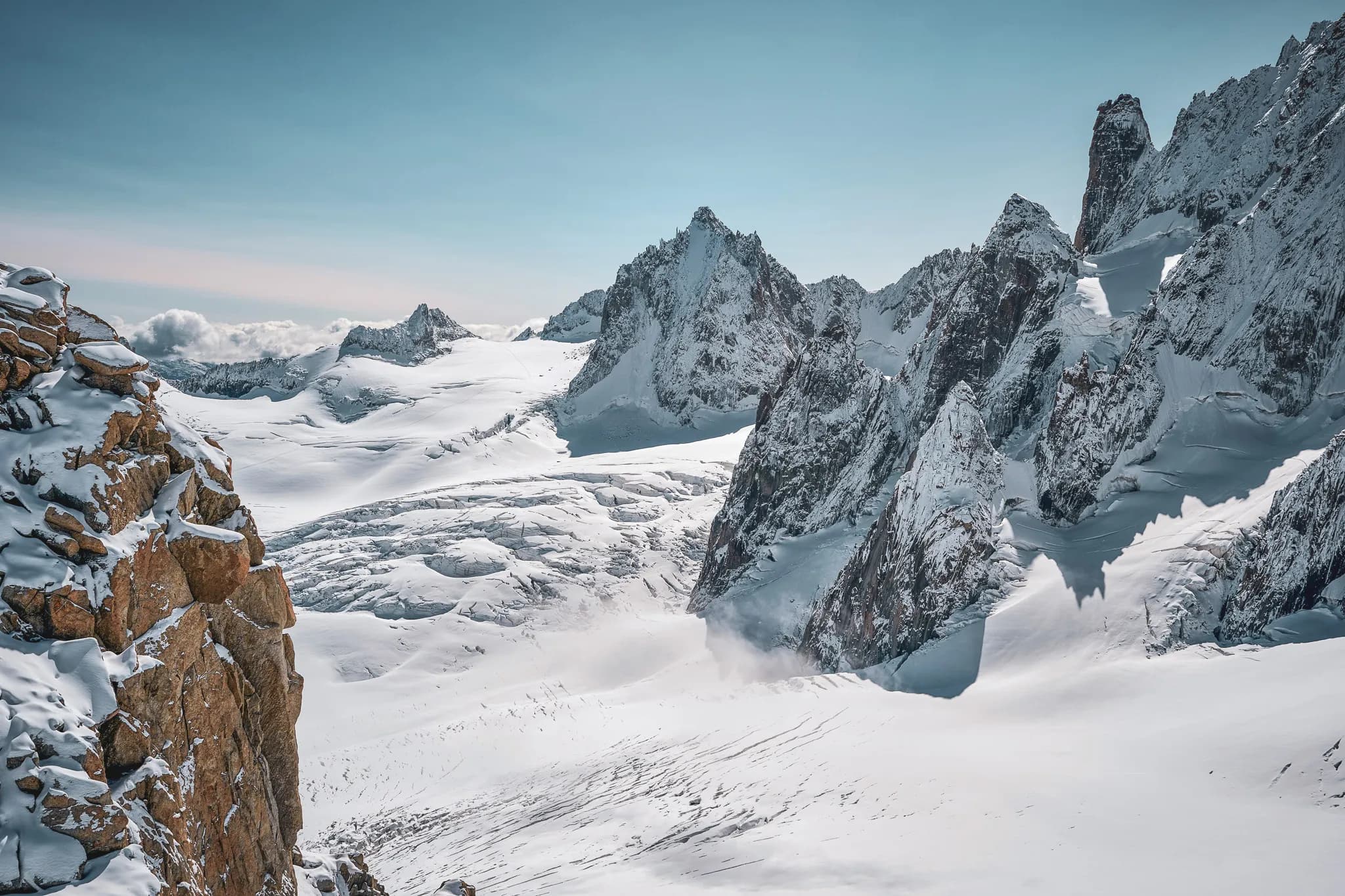 Un paysage montagneux avec des sommets majestueux recouverts de neige. Des falaises rocheuses se dressent sur le côté, tandis qu'un vaste glacier et un terrain enneigé s'étendent à perte de vue. Le ciel est