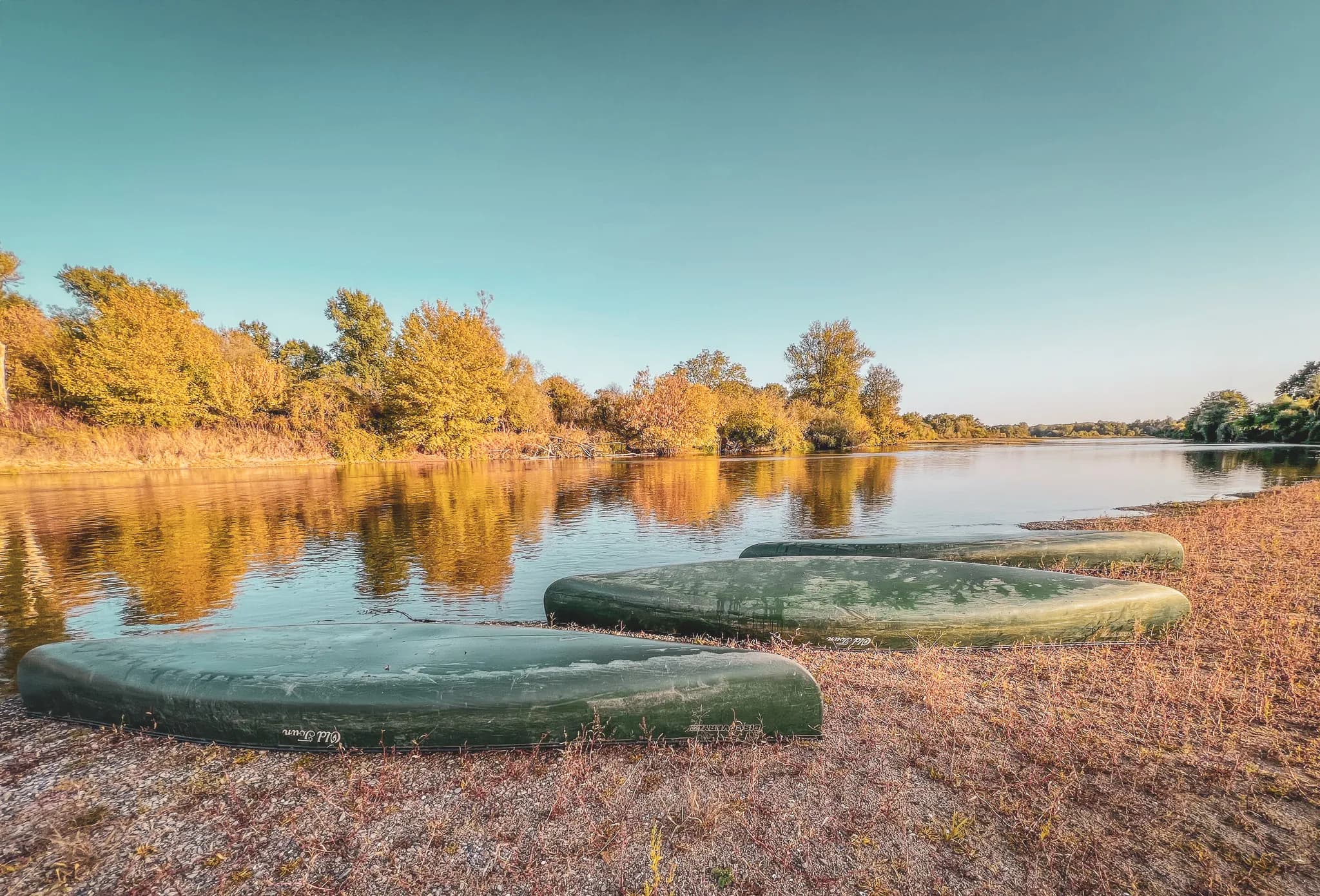 A tranquil riverside scene, with several kayaks overturned on the shore. The kayaks are green in colour and are positioned on a bed of gravel and grass. In the background, trees