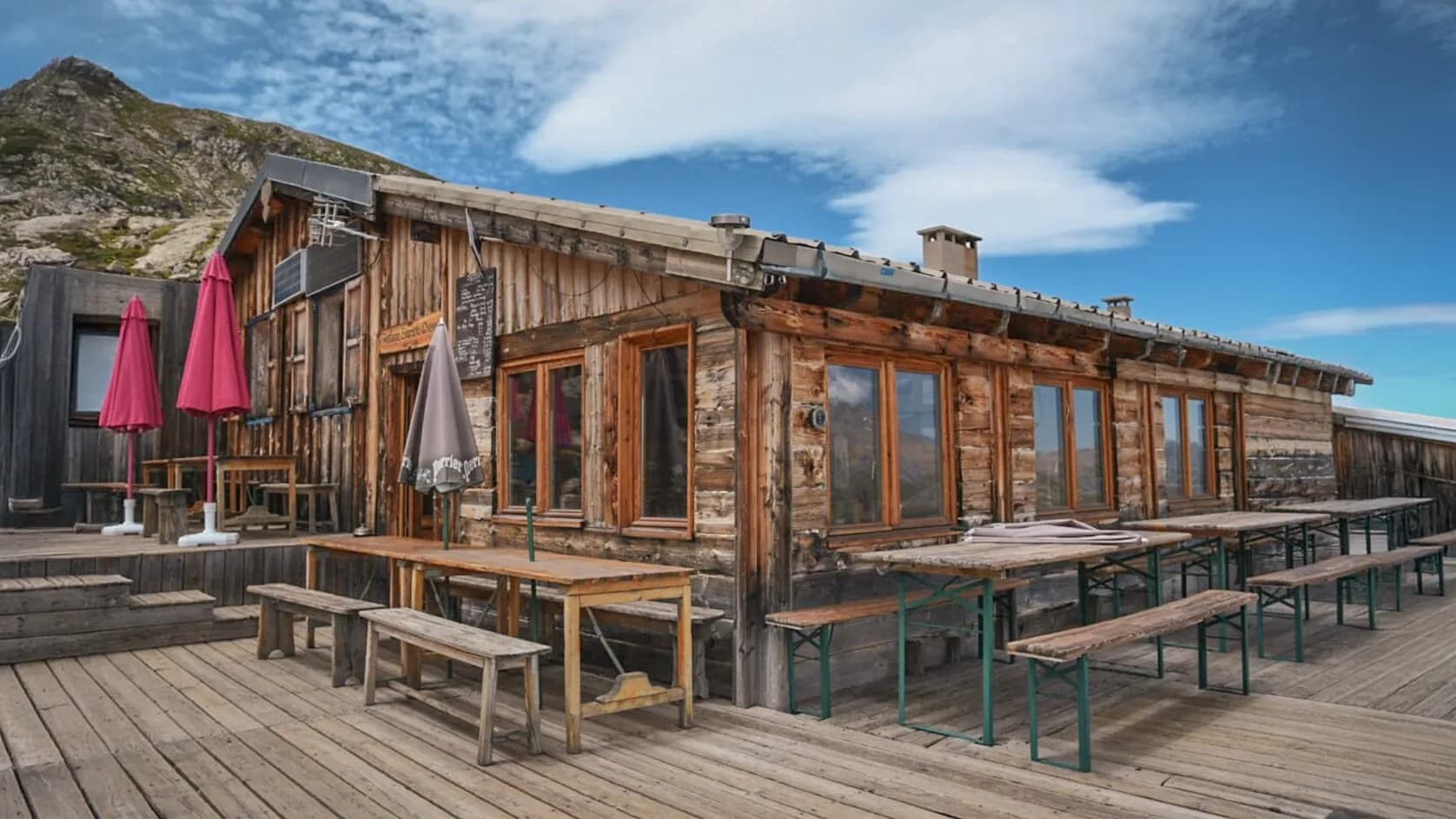 A wooden chalet in the mountains, with a wooden terrace and several tables and benches outside. Two red parasols stand in front of the entrance, adding a touch of colour. Wooden windows reveal the interior