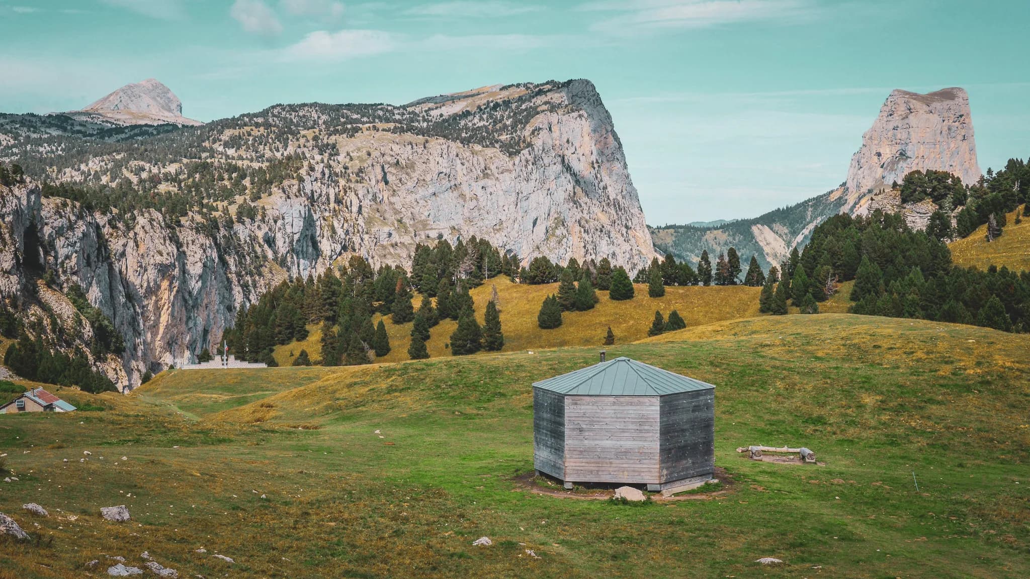 An octagonal wooden shelter stands on a vast grassy area surrounded by majestic mountains. The green hills are dotted with conifers, with green cliffs in the background.