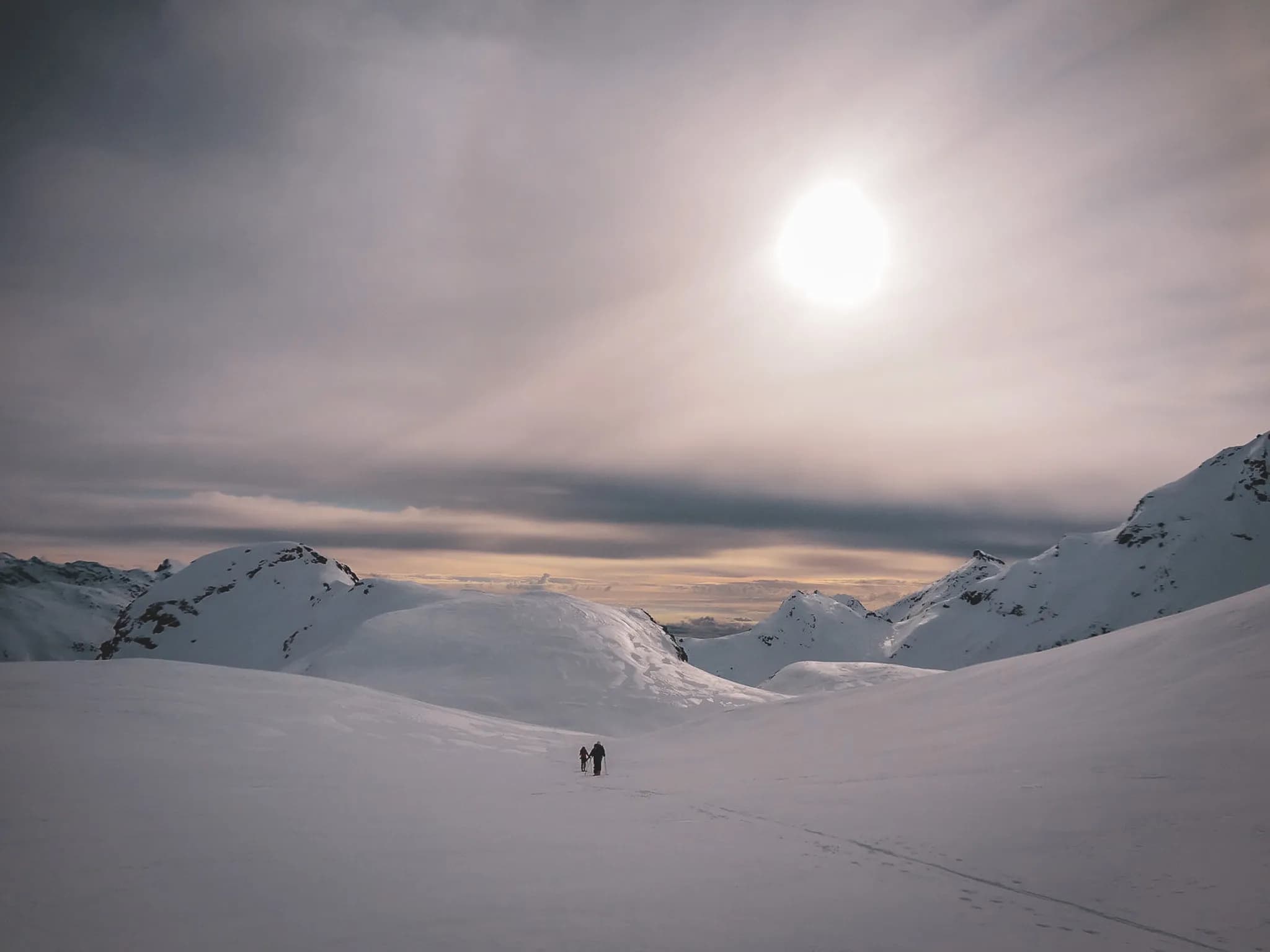 A snowy landscape, with snow-capped mountains looming on the horizon beneath a cloudy sky. The sun shines faintly, creating a soft, peaceful atmosphere. Two hikes