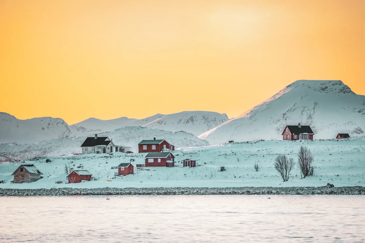 A winter landscape with colourful houses, mainly red, clustered on a snow-covered hill. In the background, majestic mountains rise up under a yellow-tinted sky, indicating either sunrise or sunset.