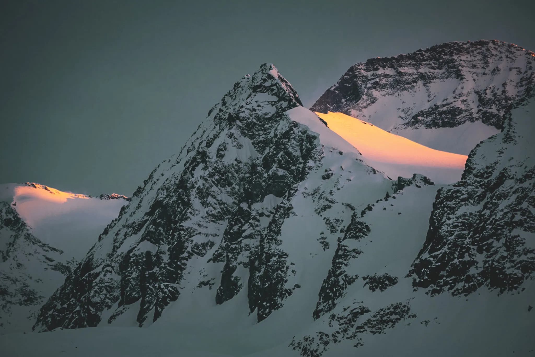 Majestic snow-capped mountains, where the peaks rise from dark, textured rock. A golden glow of sunlight reflects off the snow, creating a contrast