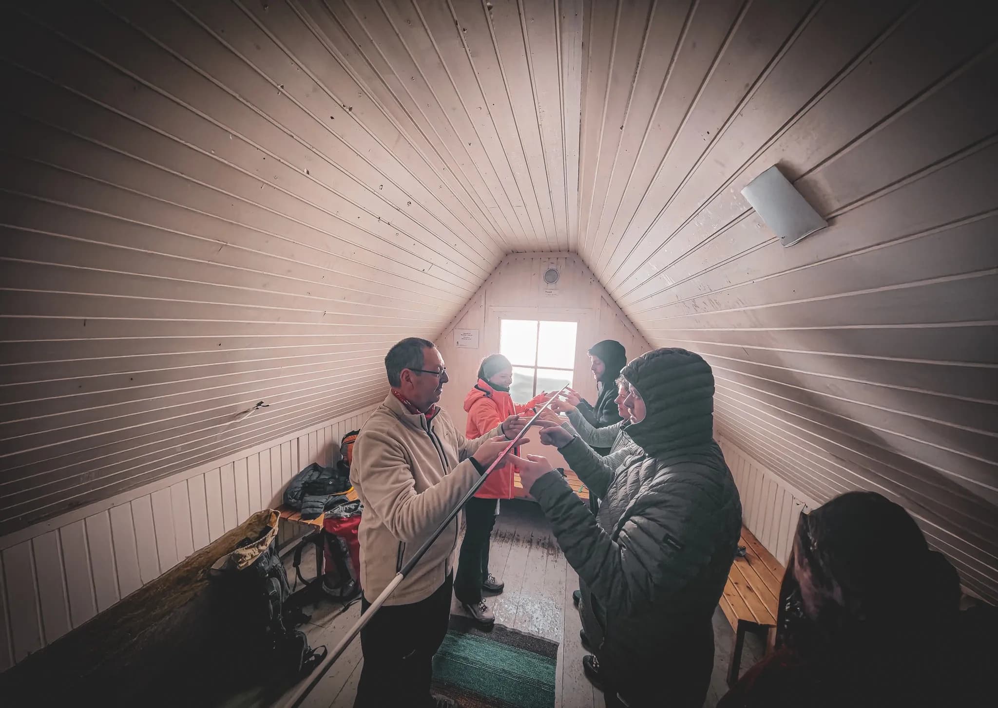 A group of four people are gathered inside a Swedish refuge, with light-coloured wooden walls and floor. They appear to be engaged in a collaborative activity, manipulating a tent peg.