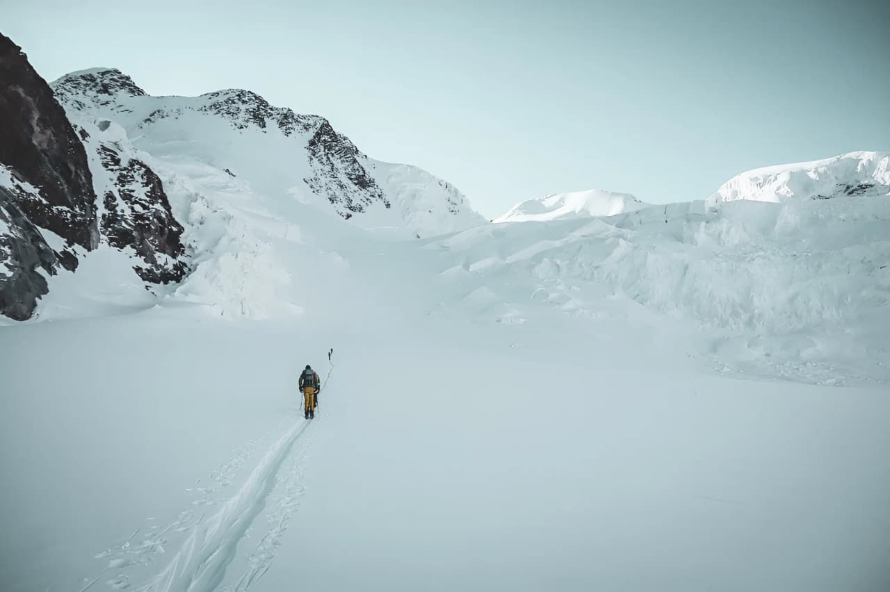 A lone skier crosses a vast glacier under a clear sky, surrounded by majestic mountains.