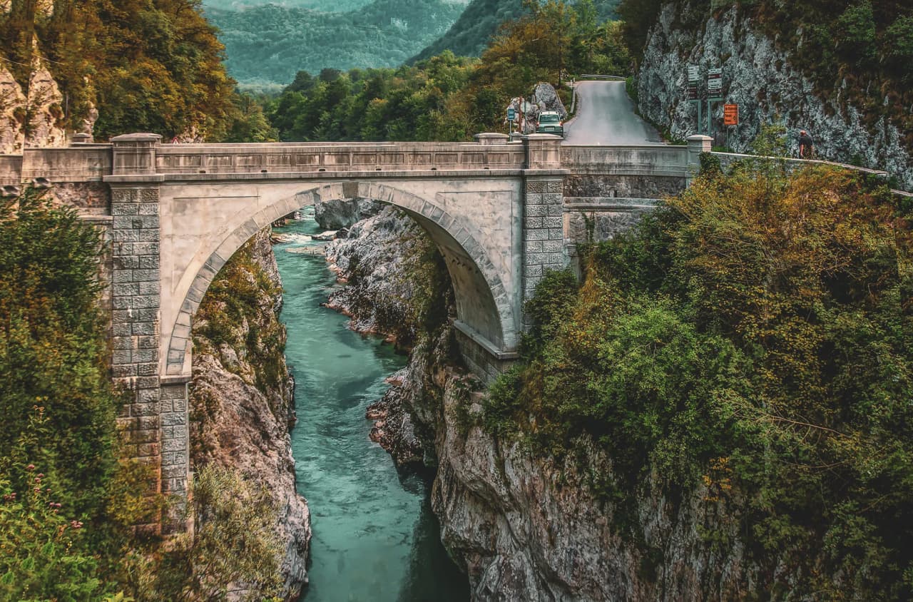 A stone bridge stretches majestically over a gorge, overlooking a brilliant turquoise river. Steep rock faces line the river, while a winding road looms ahead.
