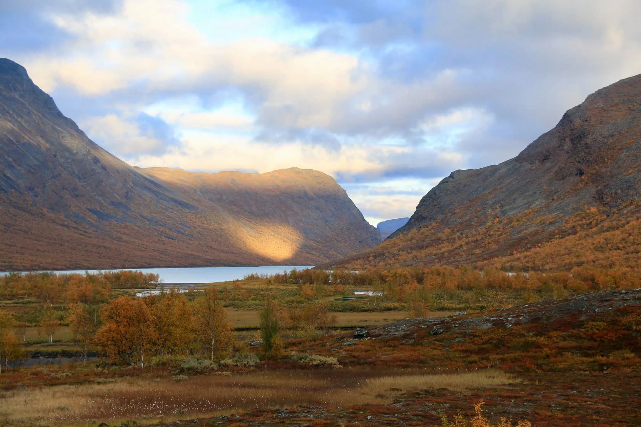 A majestic natural landscape with mountains framing a valley. The sunlight plays with the clouds, creating soft shadows on the rocky slopes. In the background, a peaceful lake reflects the bare sky.