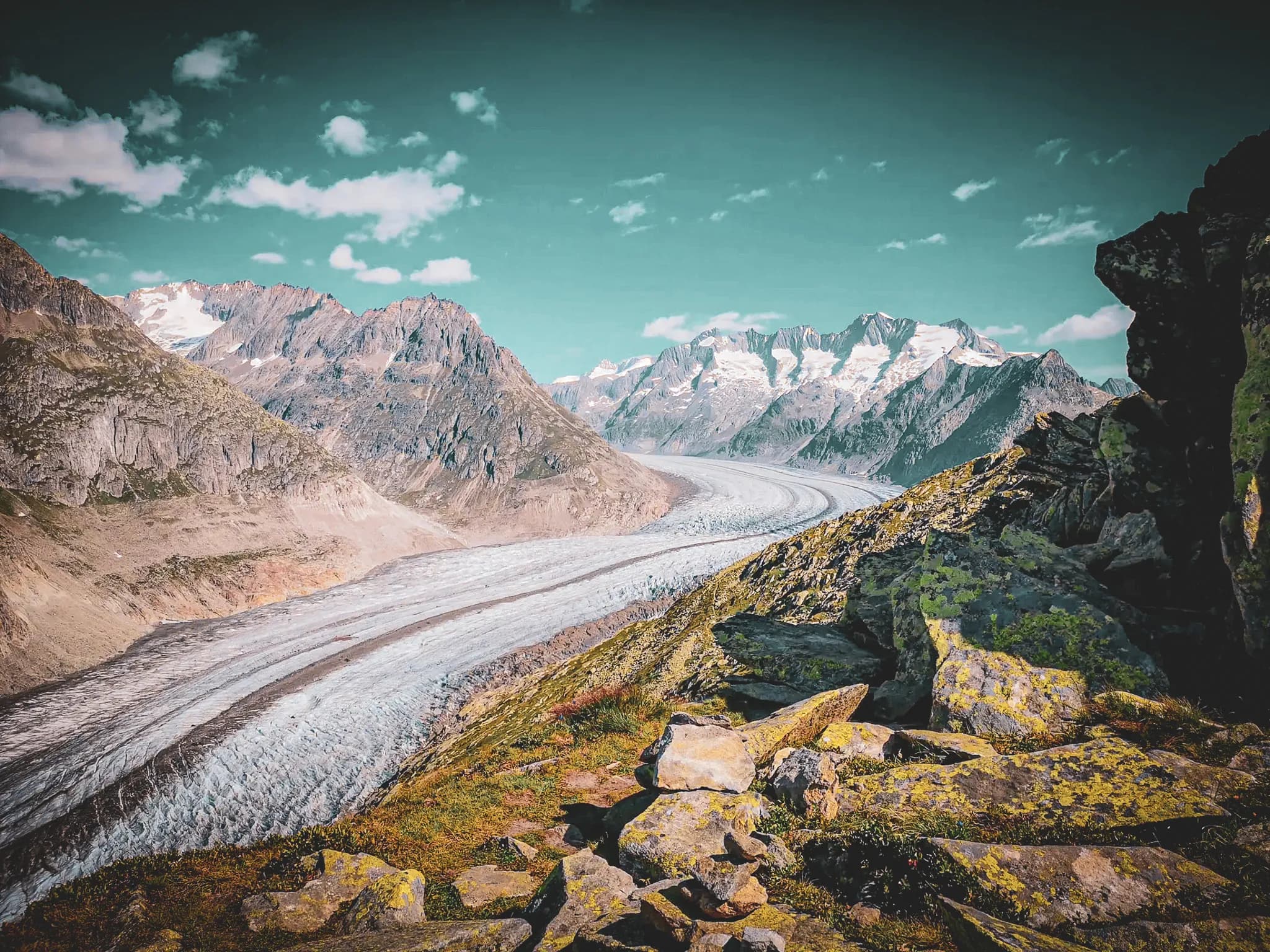 A glacier winds its way through an impressive mountain landscape, surrounded by majestic snow-capped peaks. The sky is a clear blue, dotted with white clouds.