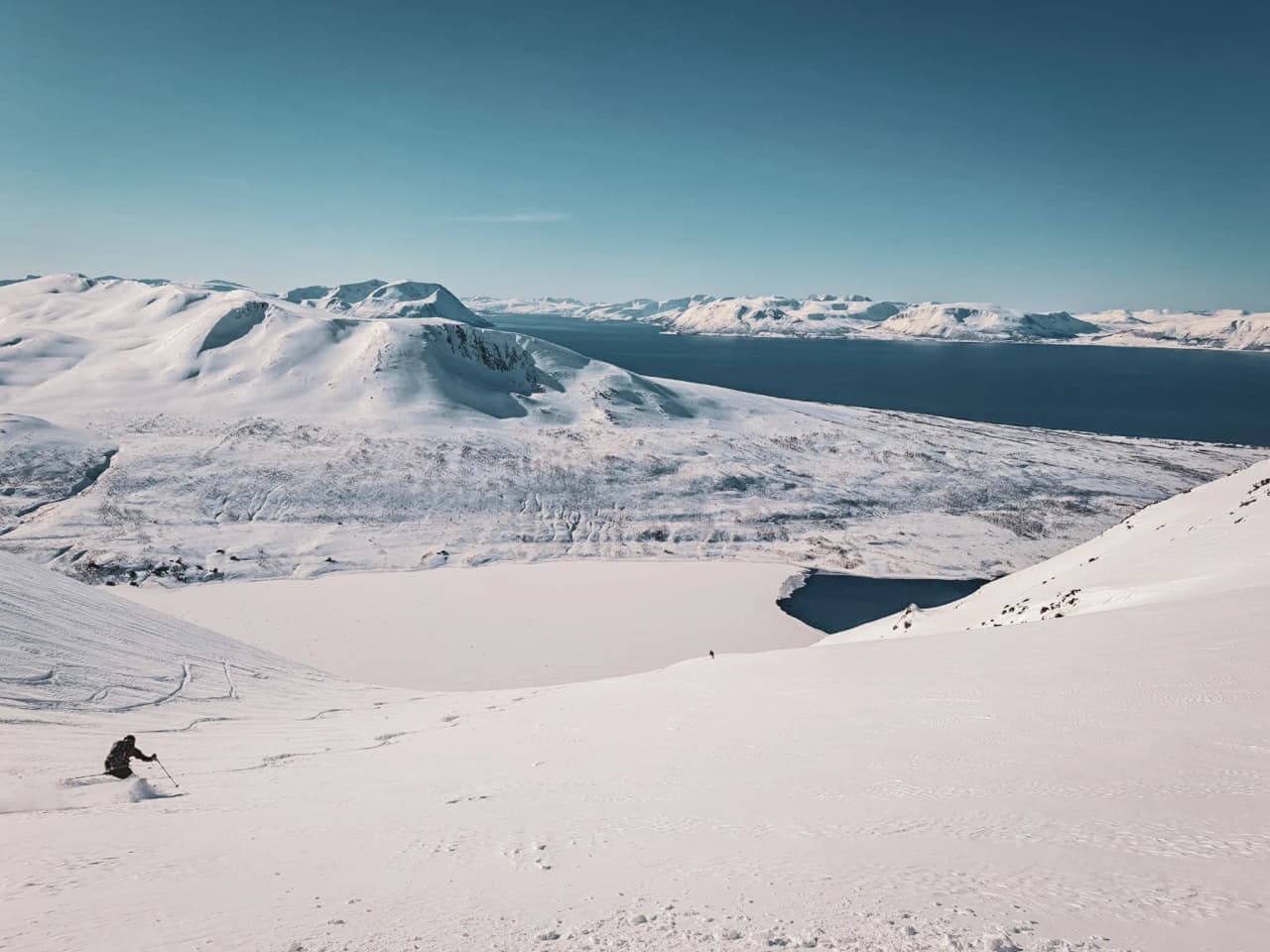 Snow-covered mountains stretch as far as the eye can see, offering a majestic winter landscape. A skier descends a snow-covered slope, leaving tracks behind him. In the distance, craggy peaks rise to the horizon.
