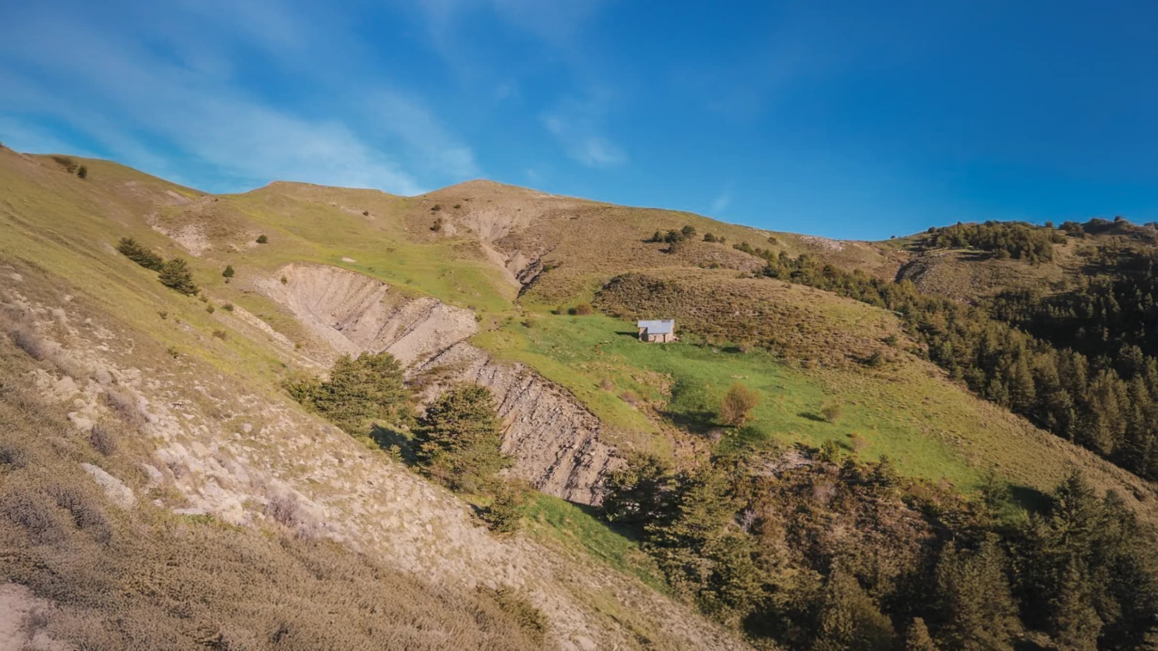 A detached wooden chalet is set on a green hill surrounded by mountains. The landscape is characterised by gentle slopes, lush vegetation and visible rock strata. The sky is clear