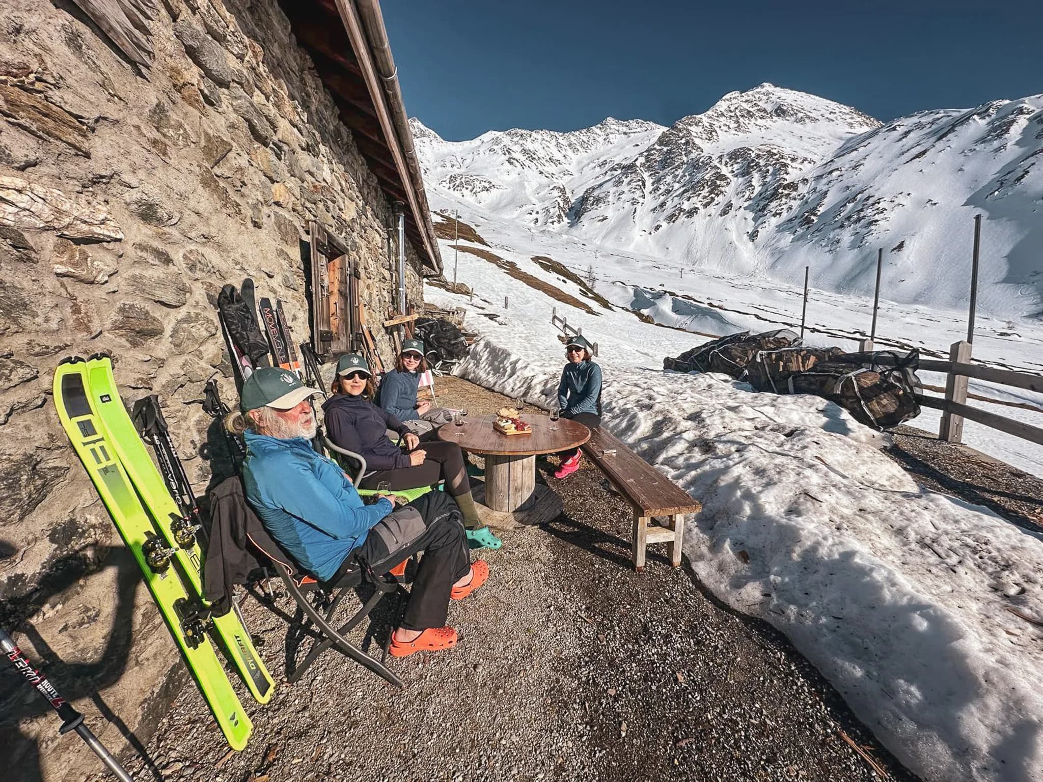 A group of hikers relax in front of a Mountain hut, surrounded by snow-capped mountains.