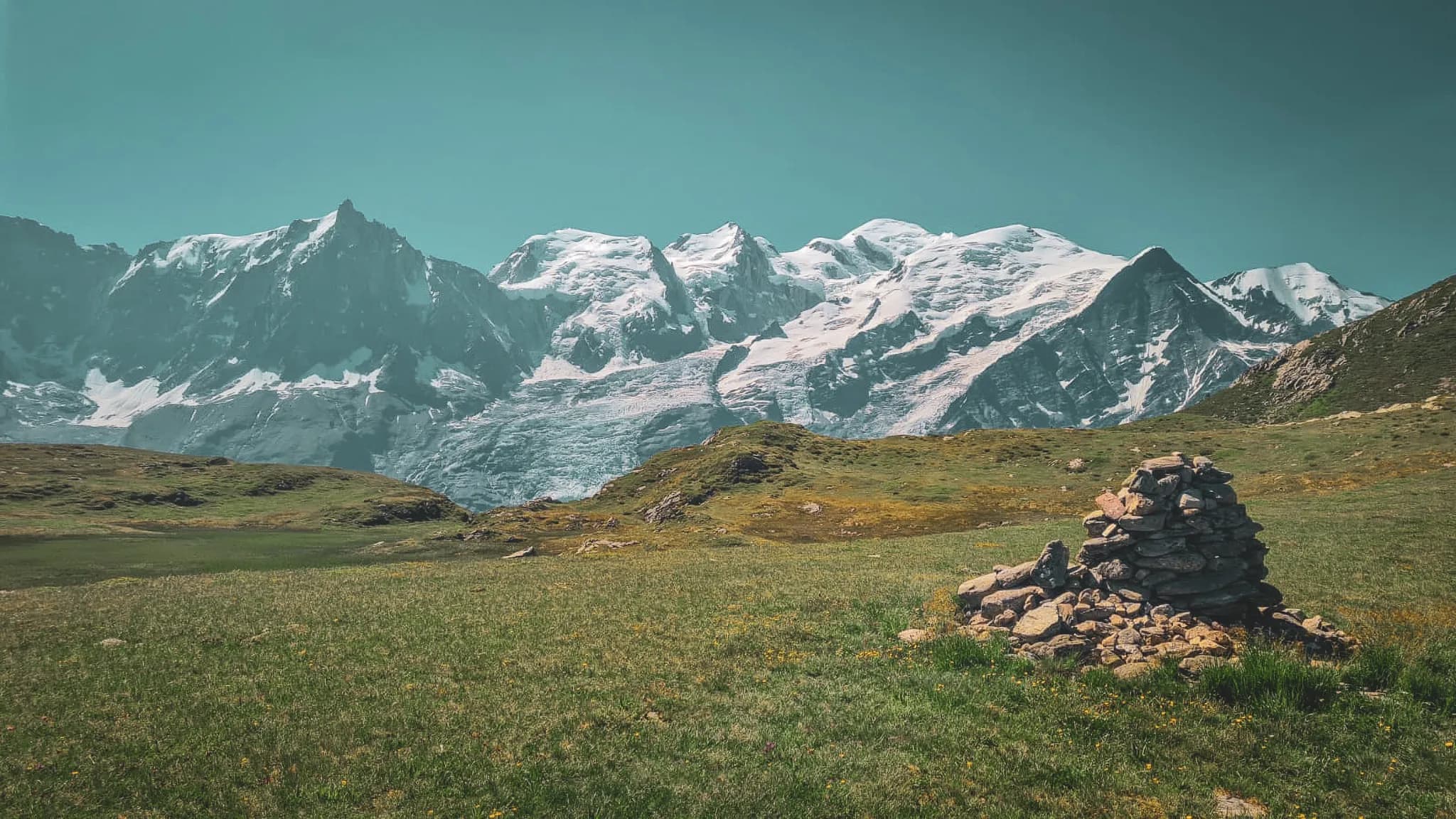 Un paysage alpin serein avec des sommets enneigés et un cairn de pierres sur une vaste prairie verte.