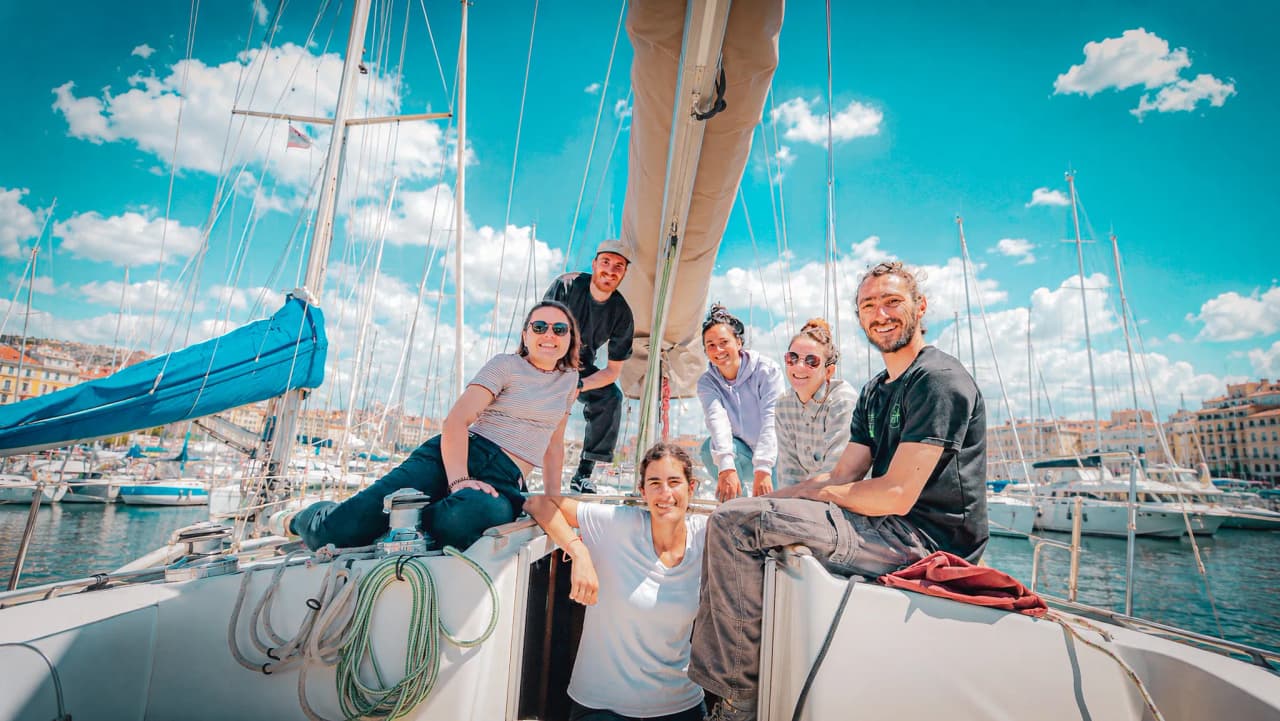 Een lachend team op een zeilboot in de Middellandse Zee, met blauwe luchten en zeilboten op de achtergrond.