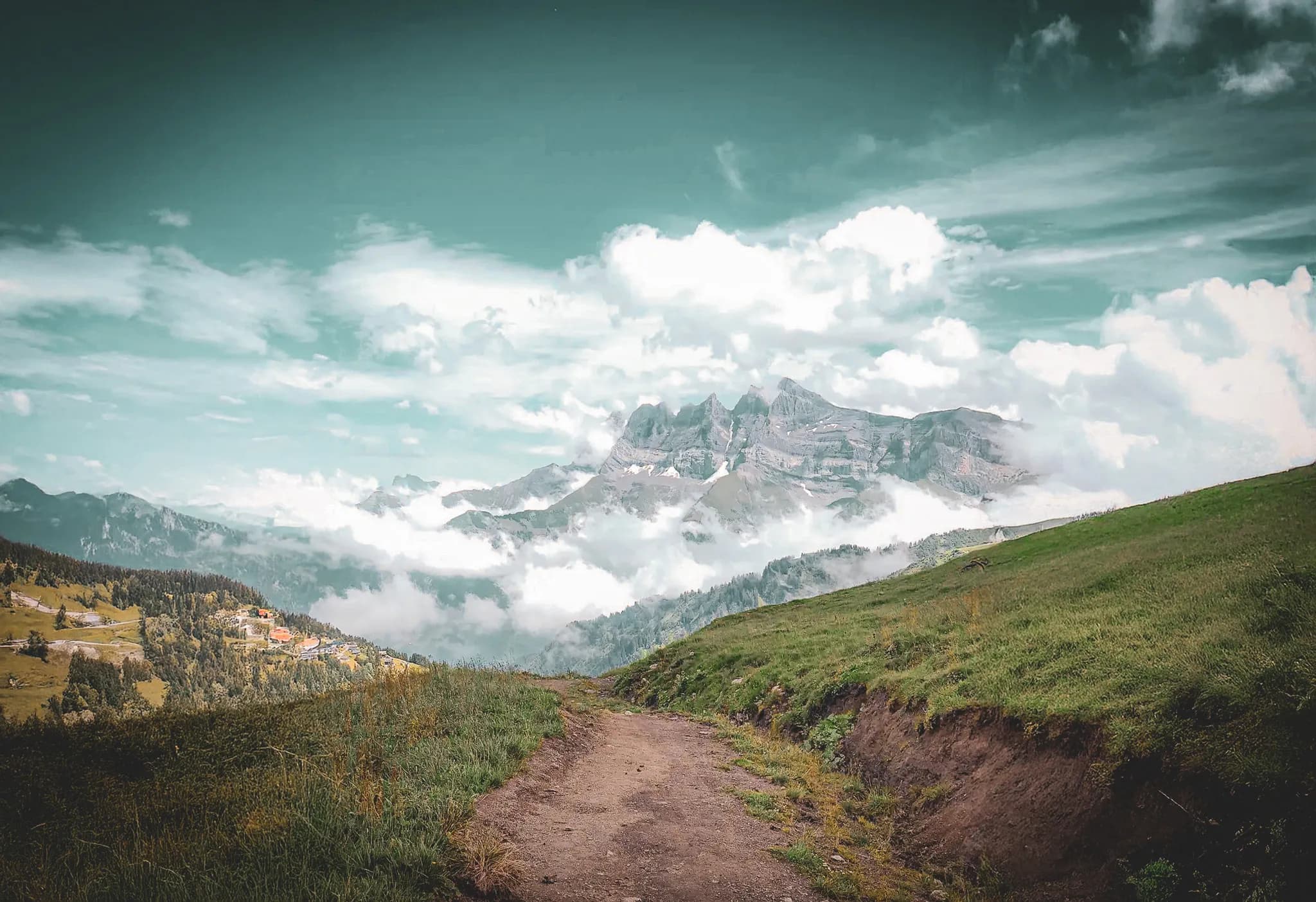 A mountainous landscape with a winding path leading through lush greenery. In the distance, steep hills are covered in light clouds, creating a mystical atmosphere. Visit