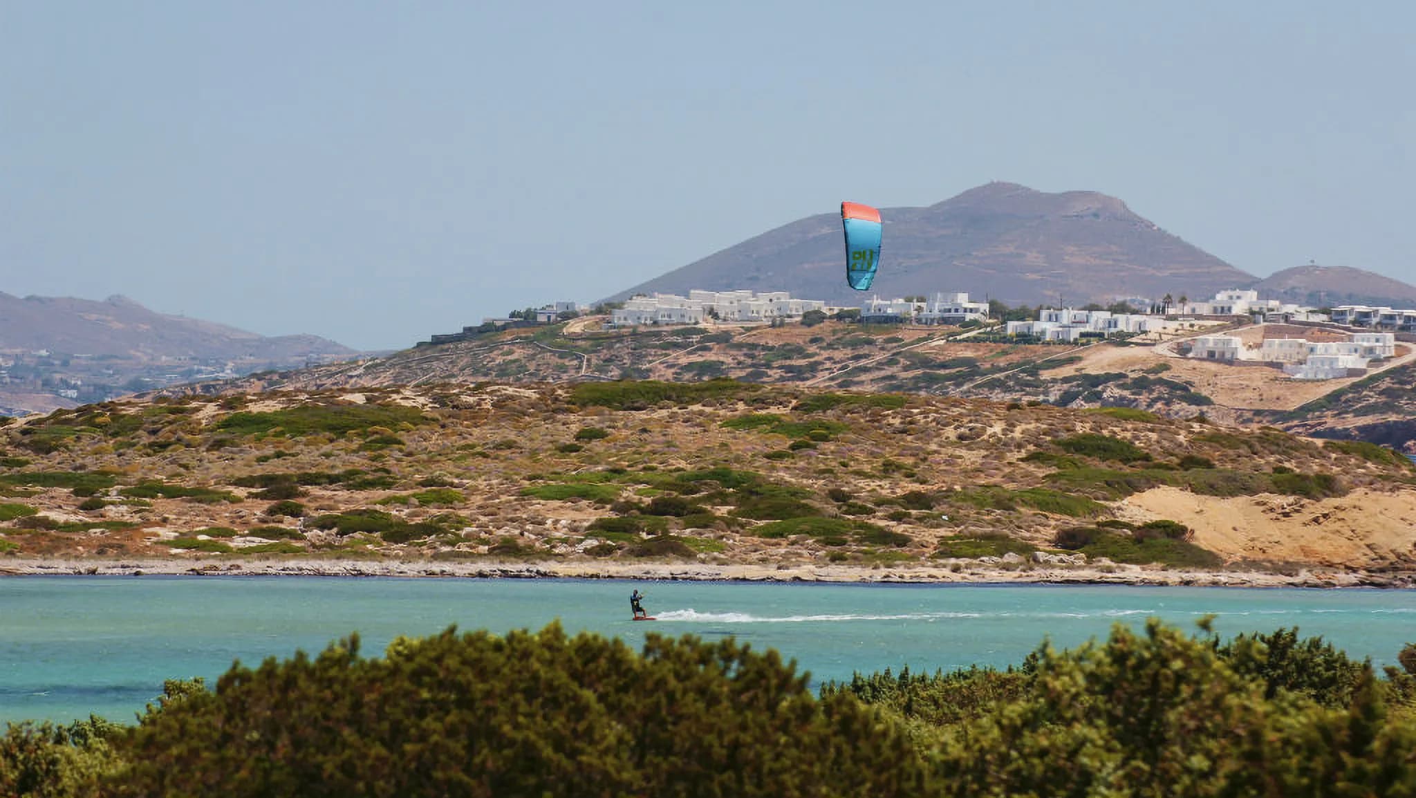 Kitesurfeur glissant sur les eaux turquoise des Cyclades, avec des collines en arrière-plan.