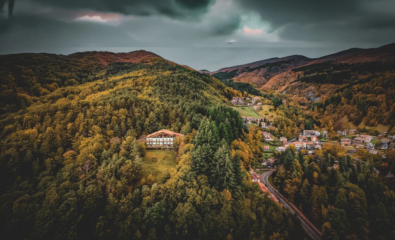 A mountainous landscape with a vast expanse of forest, mainly conifers, characterised by autumnal colours. In the centre, a white two-storey house stands on a gentle slope.