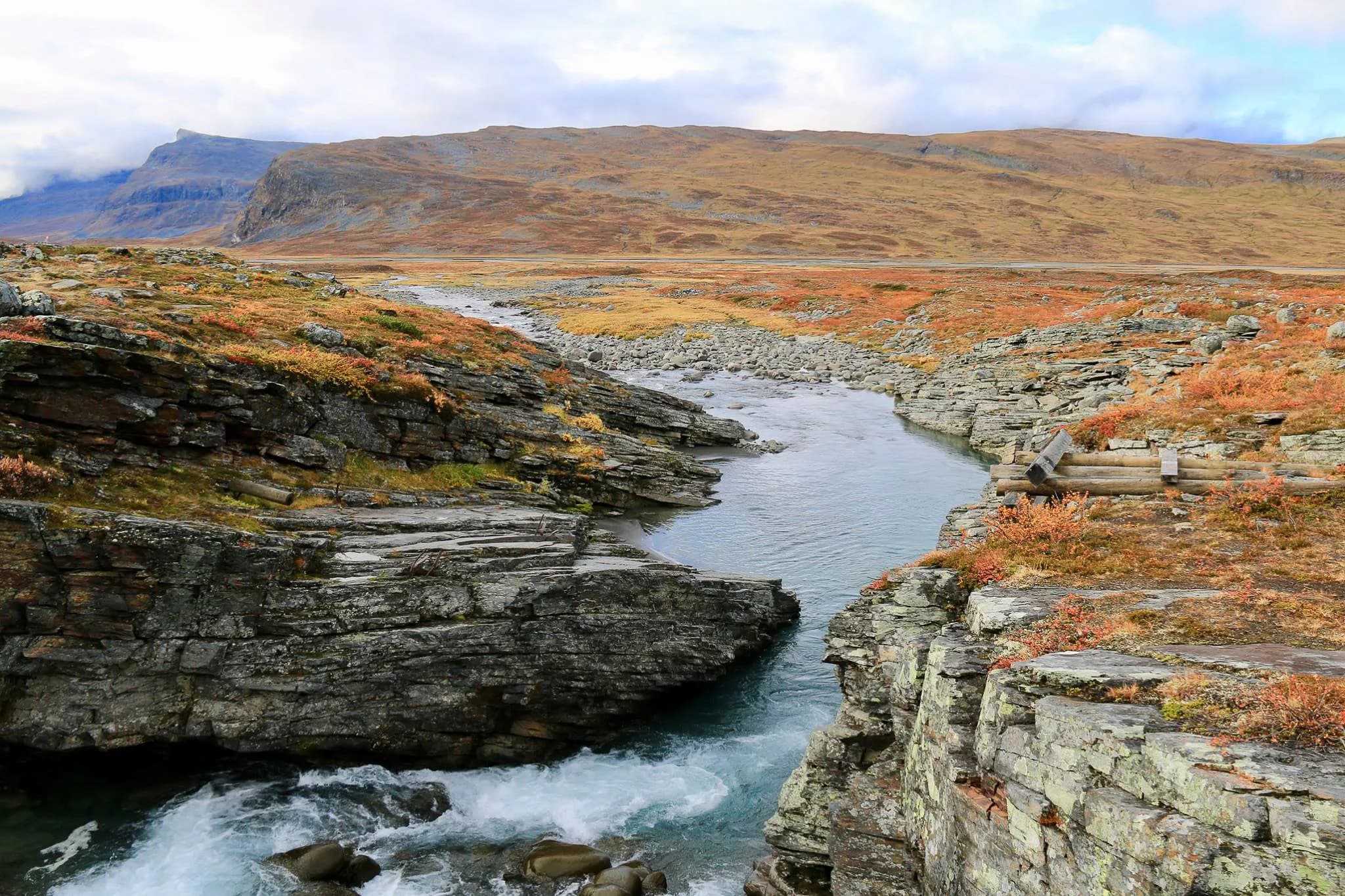 A natural landscape with a stream winding between craggy rocks. The banks are dotted with autumn foliage in shades of orange and gold. In the background, mountains offer a panoramic view.