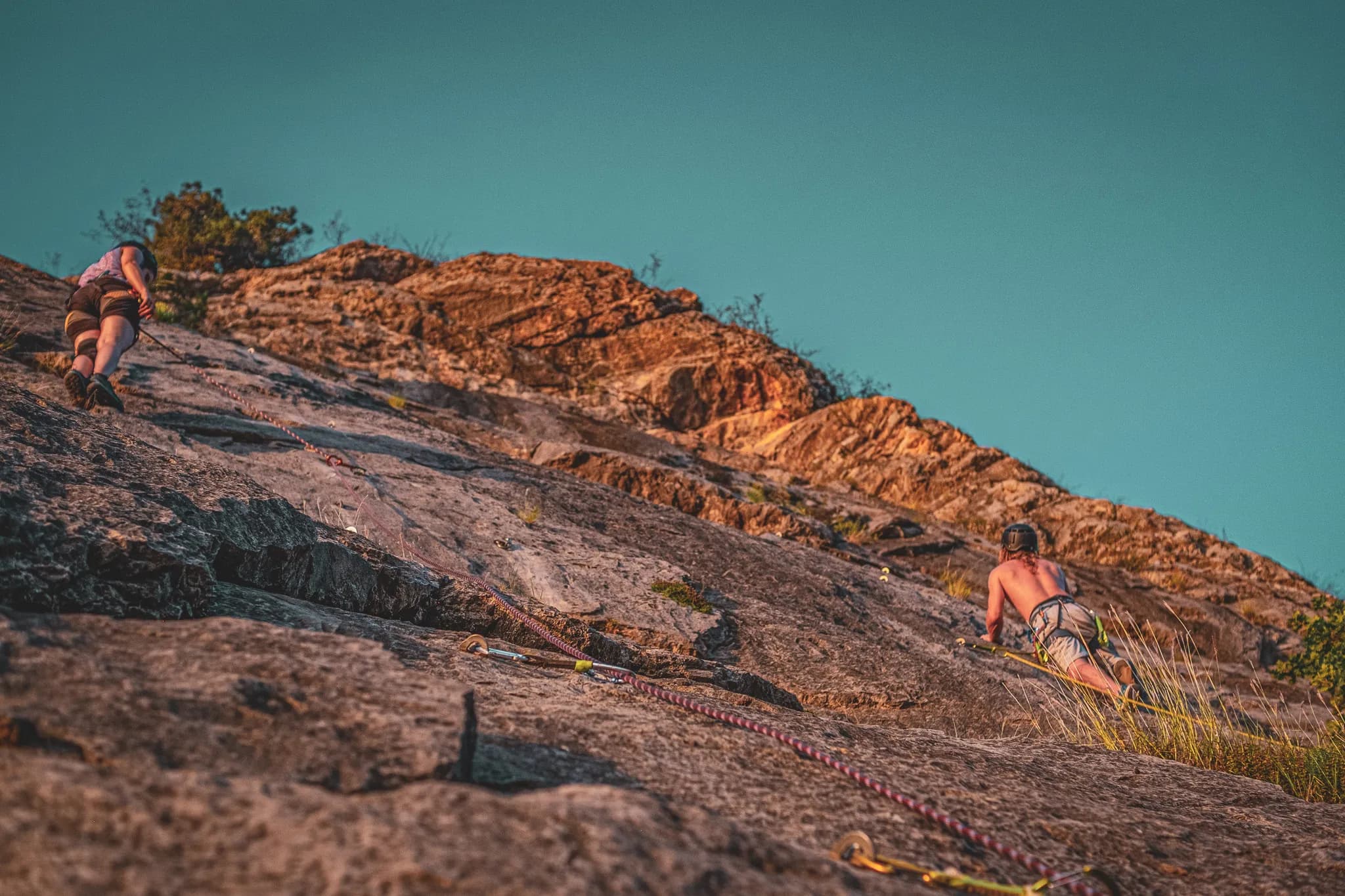 Two climbers scale a rocky cliff under a clear sky. Sunlight illuminates the rock, revealing textures and details on the surface. The climbers are wearing safety equipment and are in the middle of a climb.