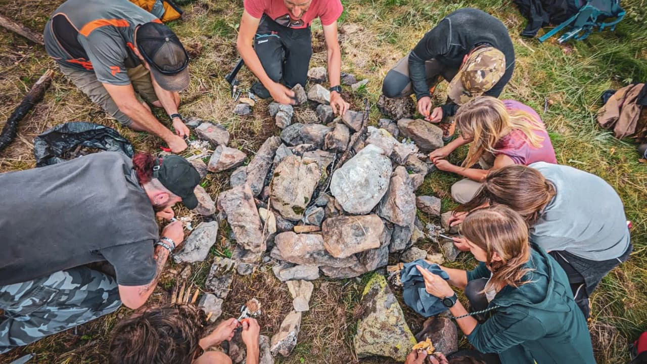 Groupe de personnes apprenant à construire un feu en pleine nature, entouré de paysages verdoyants.