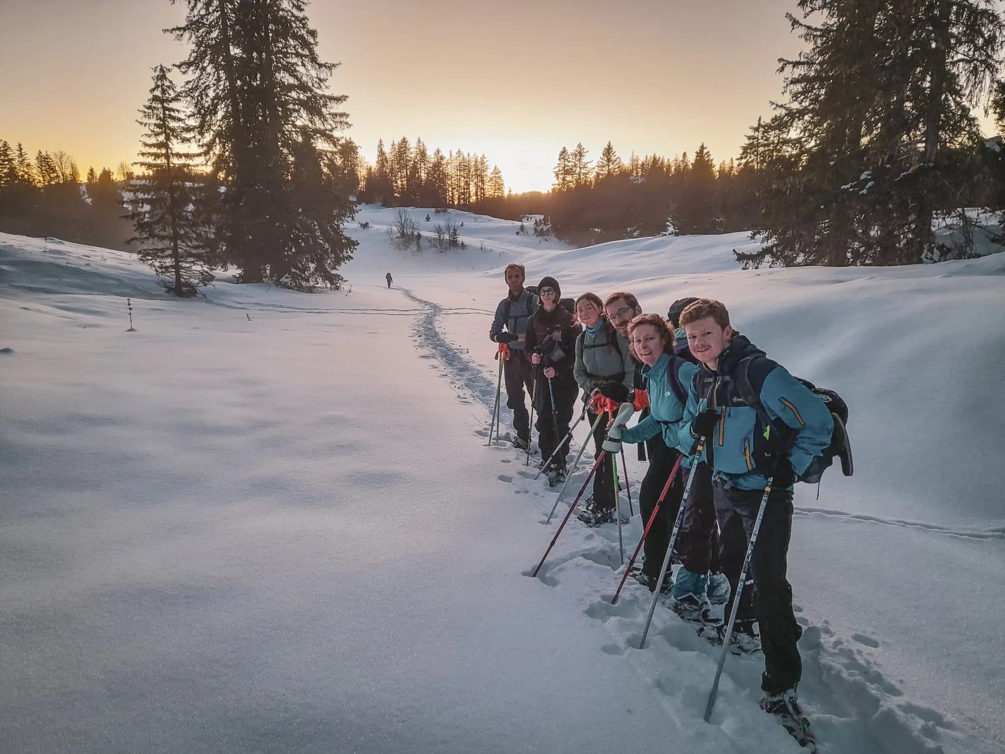 Een groep wandelaars op sneeuwschoenen doorkruist de sneeuw van de Zwitserse Jura onder een pastelkleurige hemel.