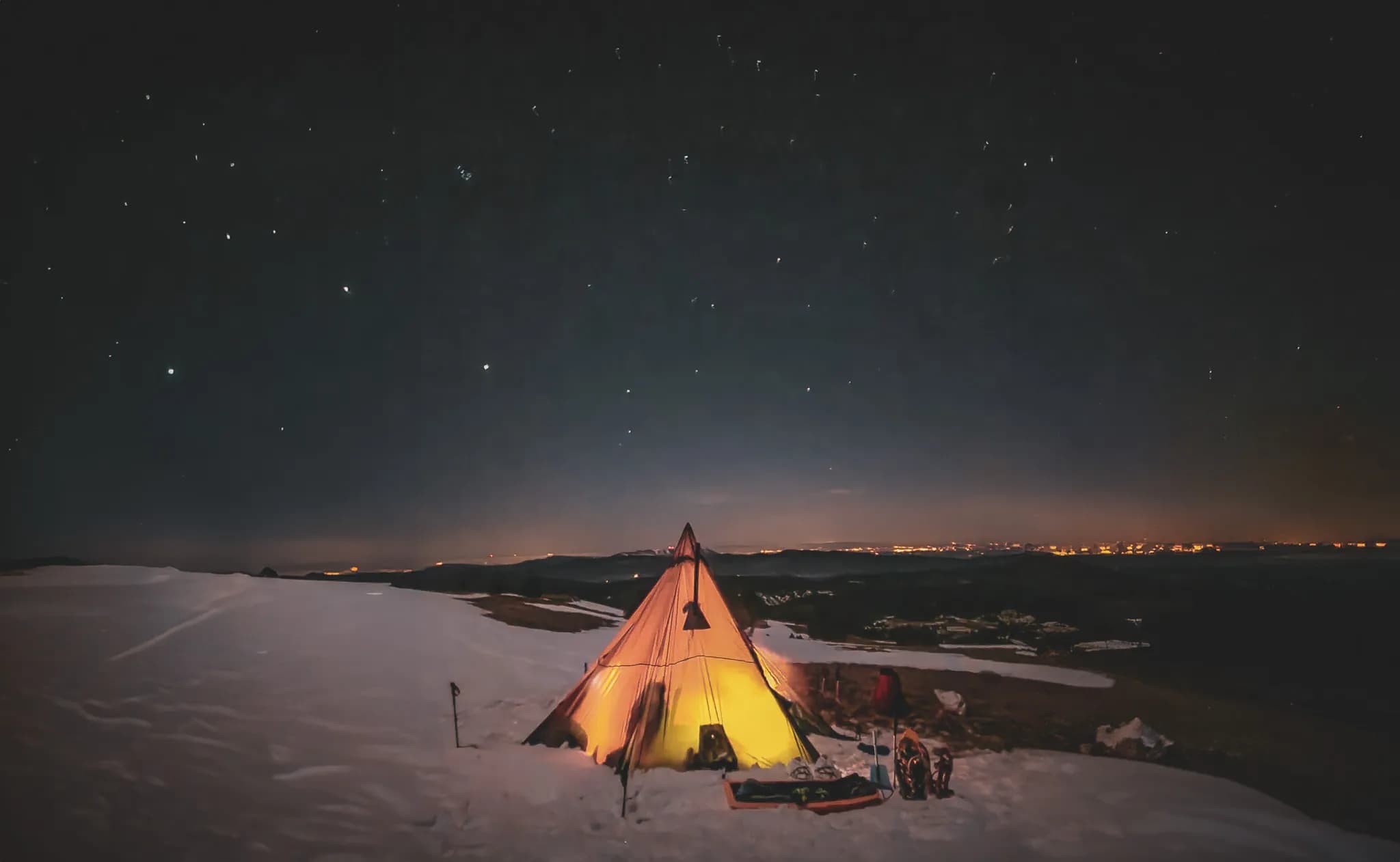 A tipi-style tent is set up on an expanse of snow, lit by soft golden light emanating from within. The surrounding landscape is mountainous, with snow formations and hills behind.