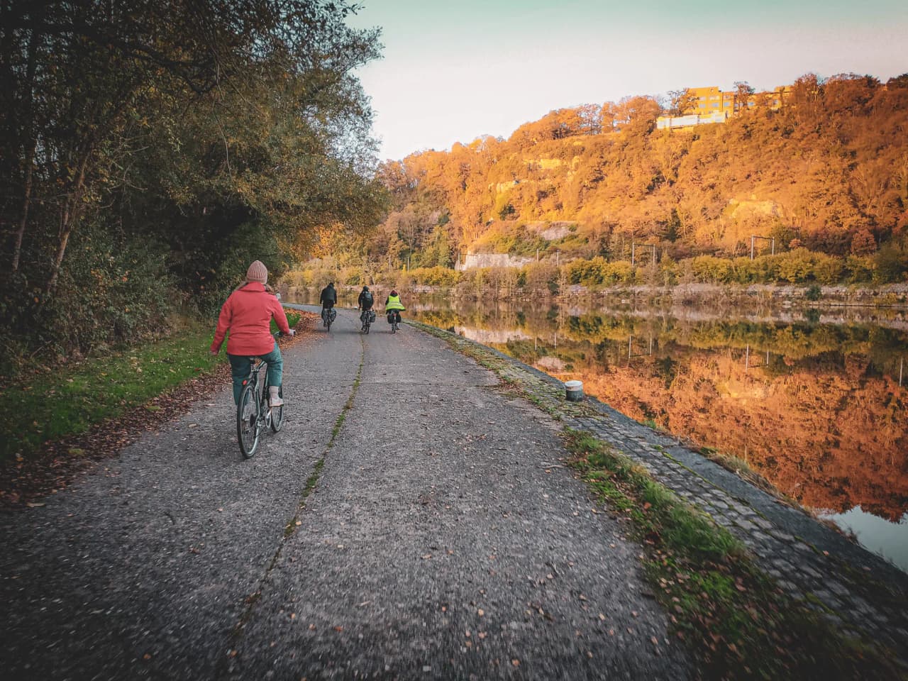 Une promenade à vélo le long d'une voie calme au bord de l'eau, entourée d'arbres aux couleurs d'automne. Un groupe de cyclistes avance sur un chemin gravillonné, tandis que l'eau reflète les nuances dor