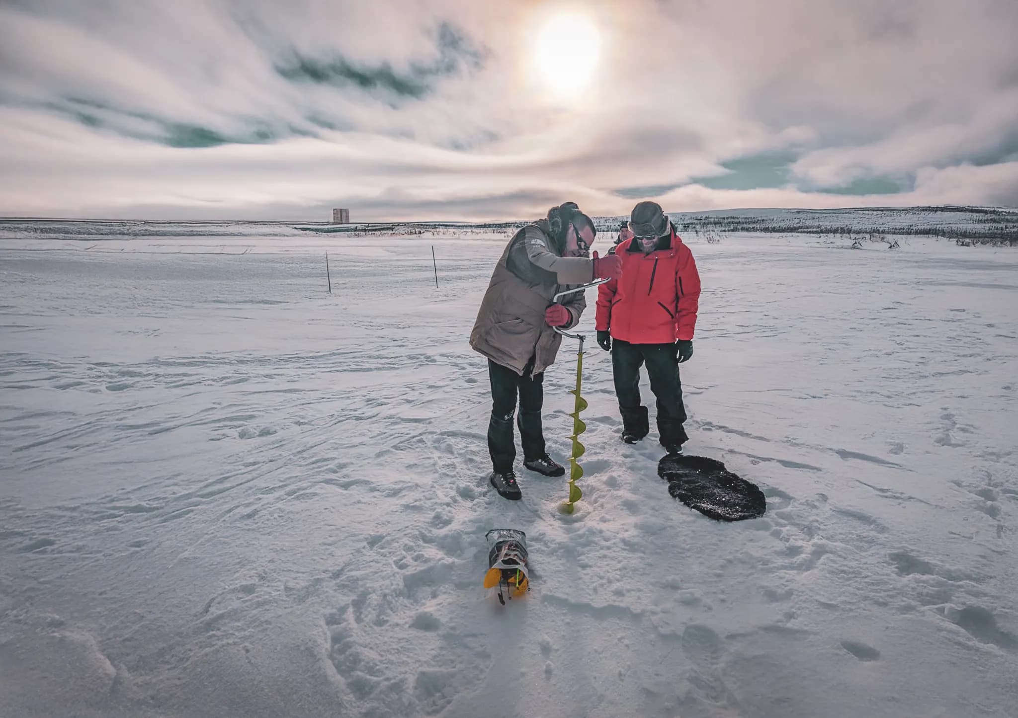 Two people are standing on a stretch of snow, with a cloudy sky in the background. One of the people, wearing a brown jacket, is using an ice drill to pierce the surface of the snow.