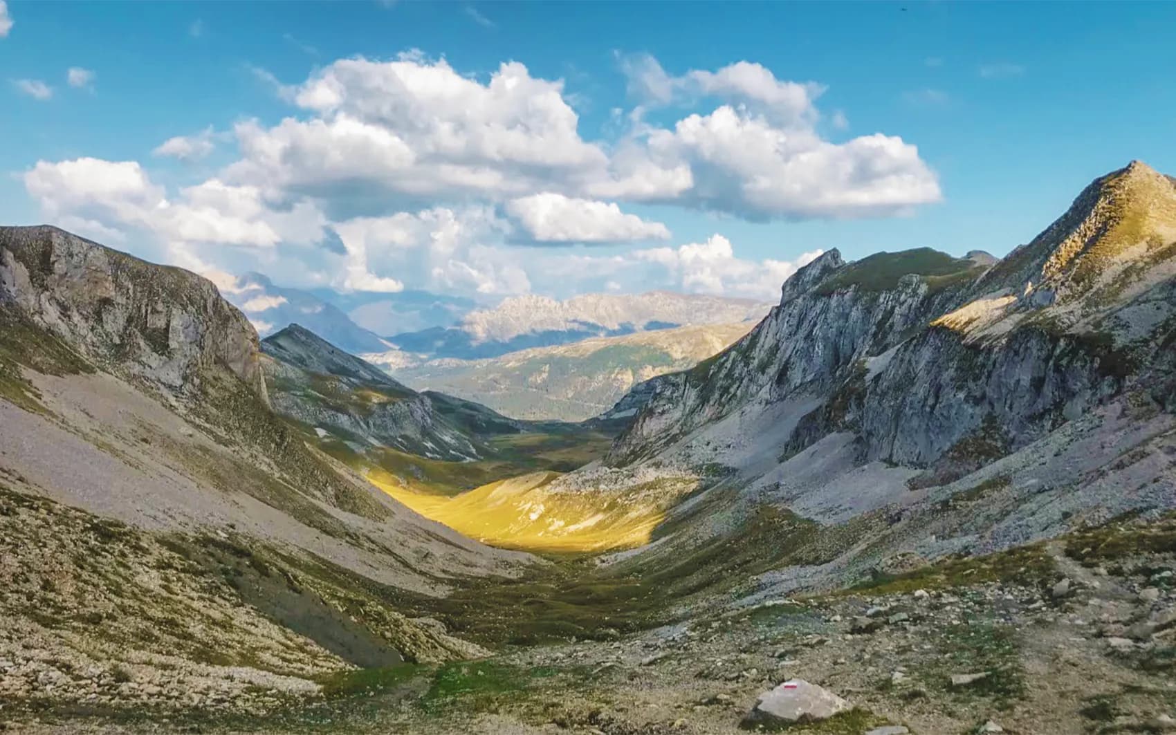 A spectacular mountain landscape with deep, wide valleys. In the foreground, rocky, grassy terrain slopes down into golden light, while the rock-covered mountainsides