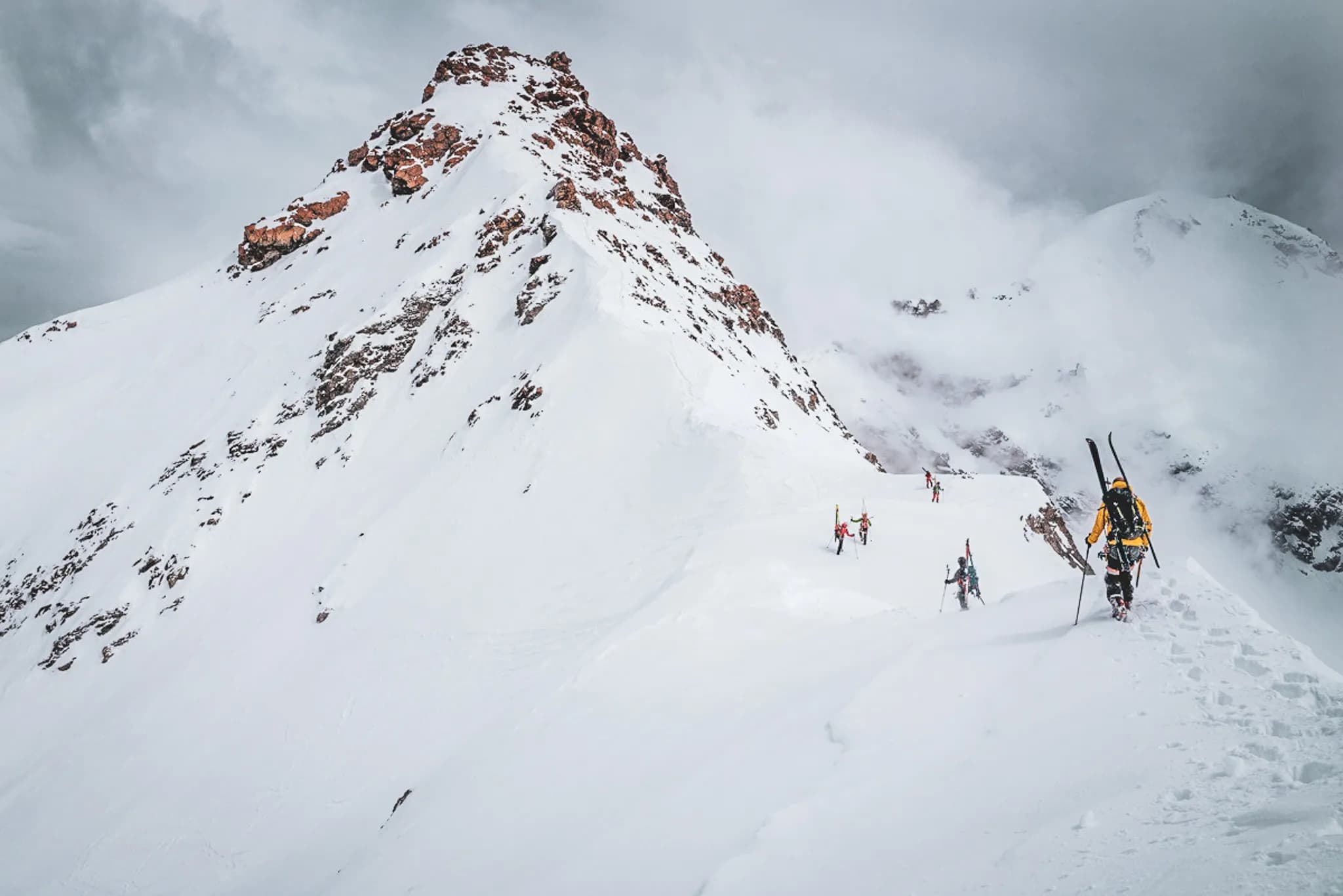 A snow-covered mountain peak, dotted with rocks, rises majestically under a cloudy sky. Skiers dressed in colourful outfits make their way cautiously up the slope,