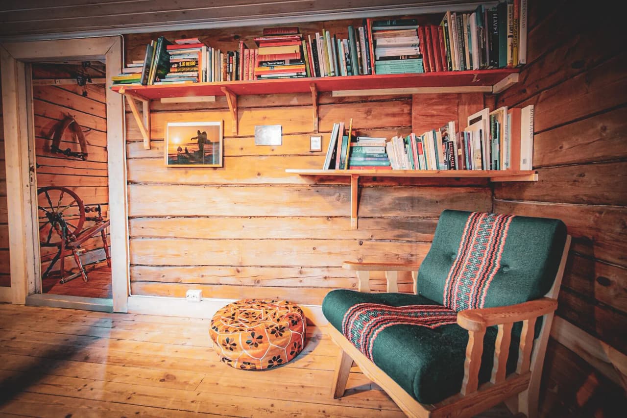 A cosy living space with a wooden wall, decorated with several shelves filled with colourful books. On one of the shelves is a photo frame showing silhouettes at the water's edge. To the right, an armchair