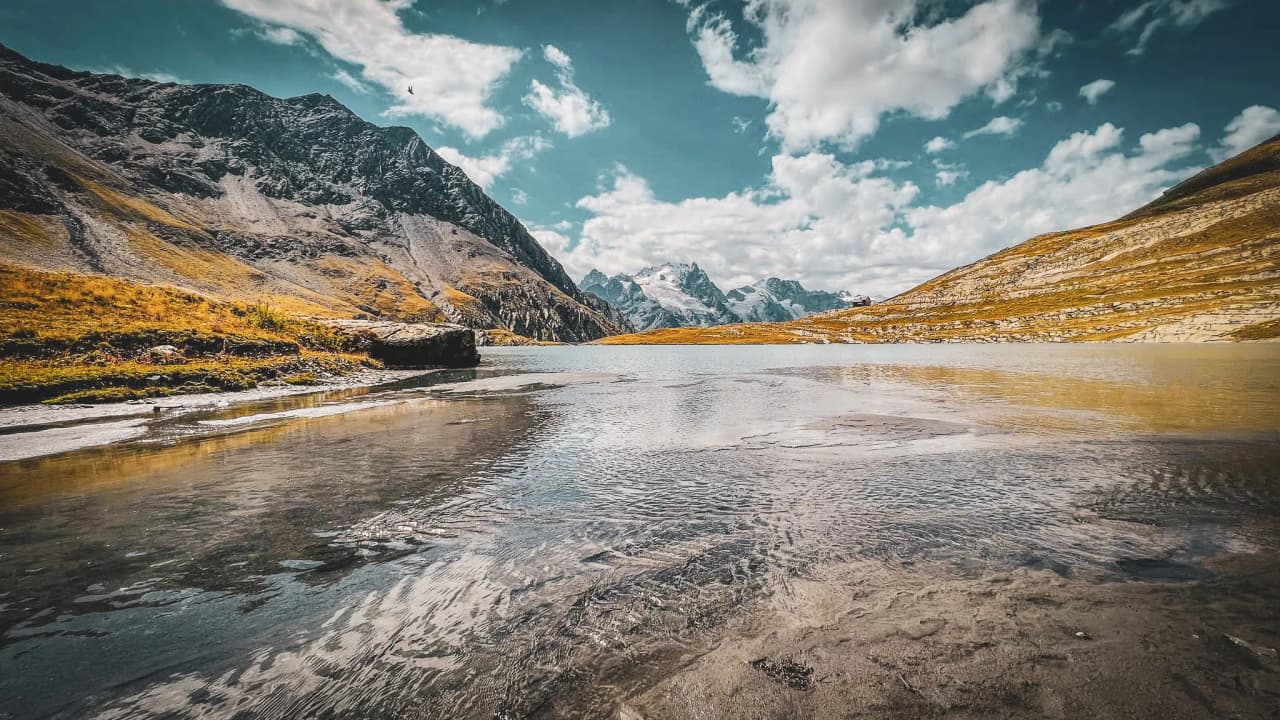Lac turquoise entouré de montagnes majestueuses, ciel bleu parsemé de nuages, invitation à l'aventure.