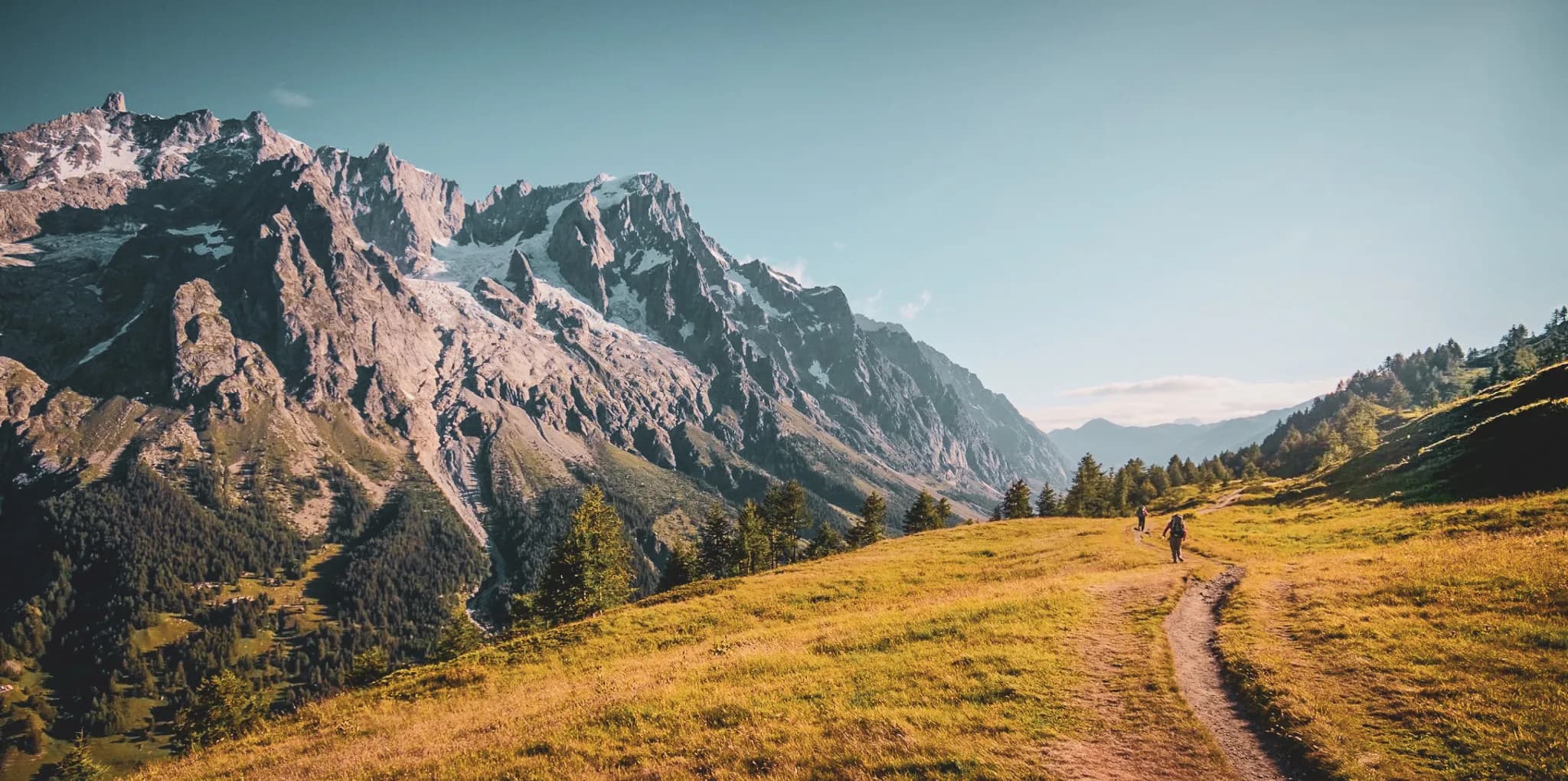 Un paysage montagneux majestueux avec des sommets rocheux recouverts de neige. Une vaste prairie dorée s'étend au premier plan, parsemée de quelques arbres. Un sentier sinueux traverse la scène, où plusieurs randon