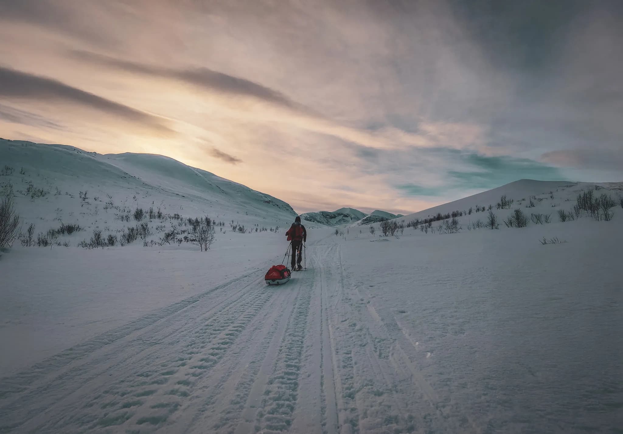 An adventurer pulls a sledge across the vast snow-covered landscape of Kingstrail in Lapland. Snow covers the land, and gentle hills stretch to the horizon. The sky is tinged with soft colours.