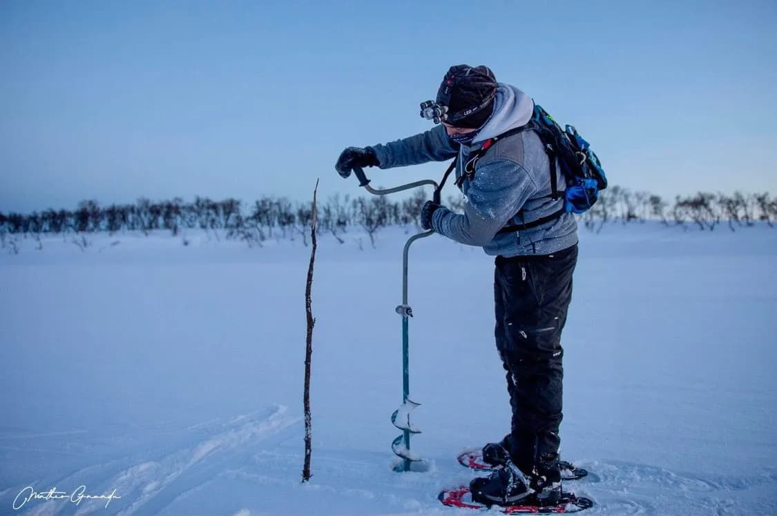 A man uses an ice drill to make a hole in the snow, wearing snowshoes and equipment adapted for winter conditions. The surrounding landscape is vast and covered in a thick layer of snow.