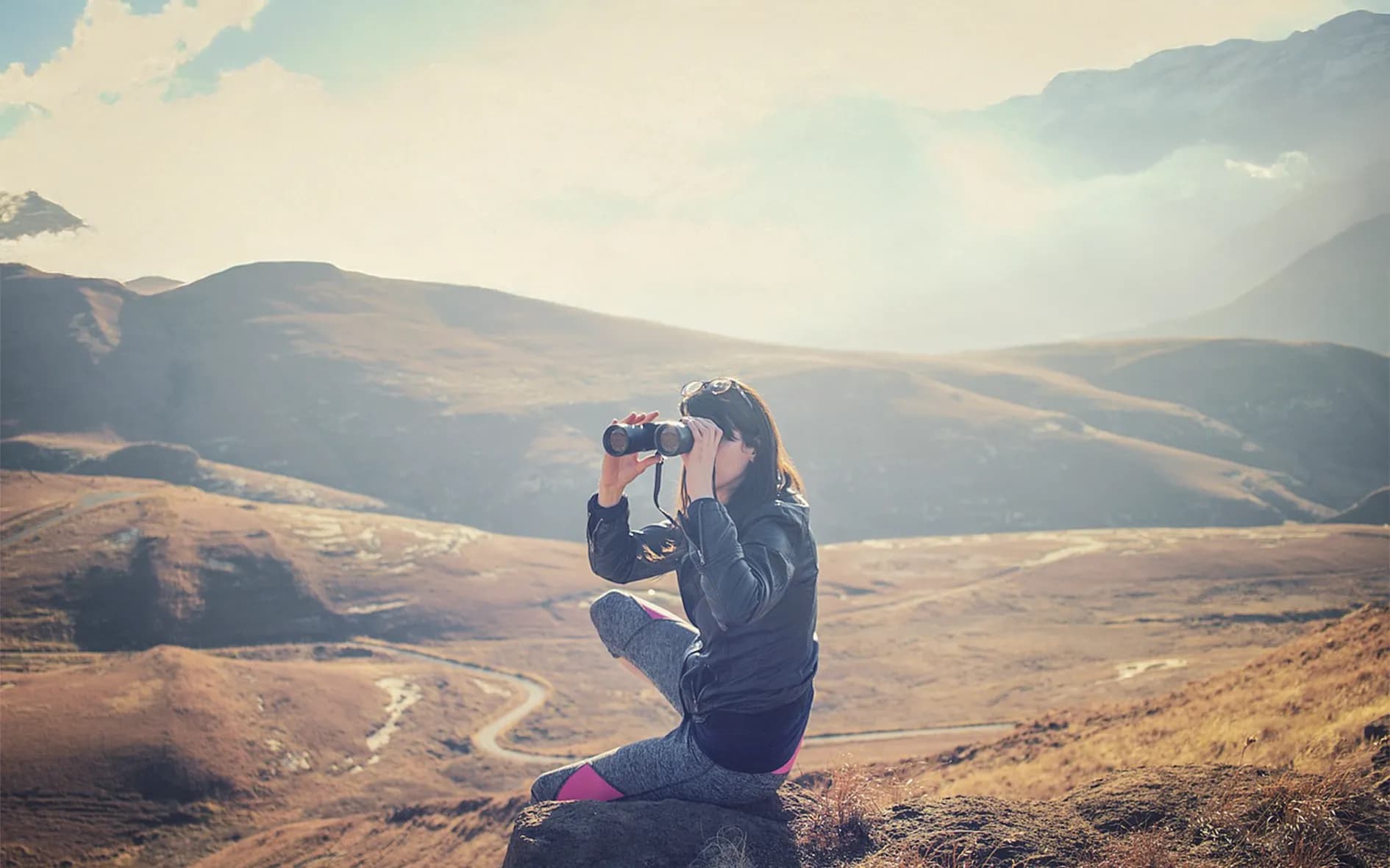 A mountainous landscape under a partly cloudy sky, with arid hills and valleys. A person is sitting on a rock, wearing a jacket and leggings, holding binoculars to observe the