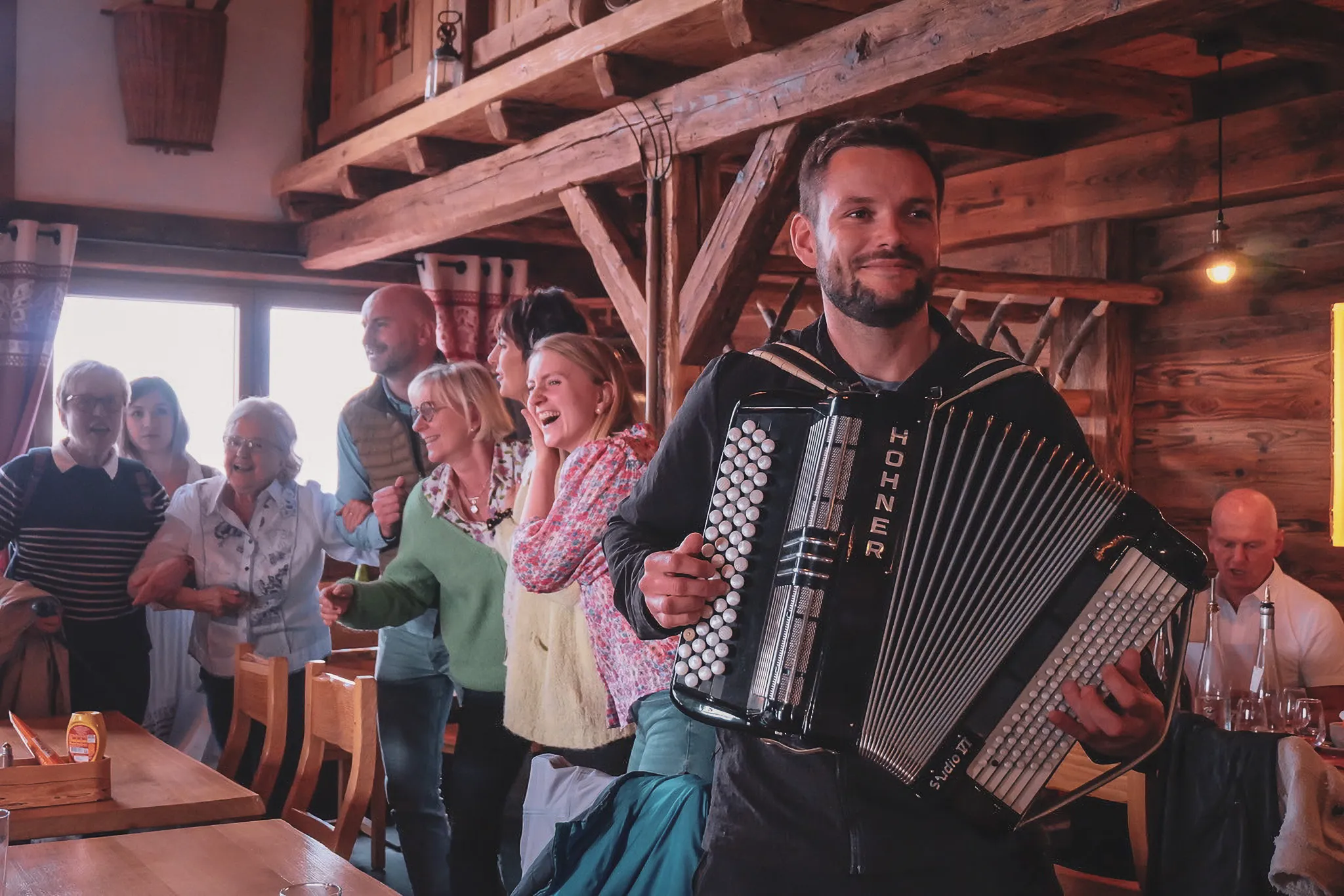 Un homme joue de l'accordéon, entouré de randonneurs joyeux dans un refuge chaleureux.