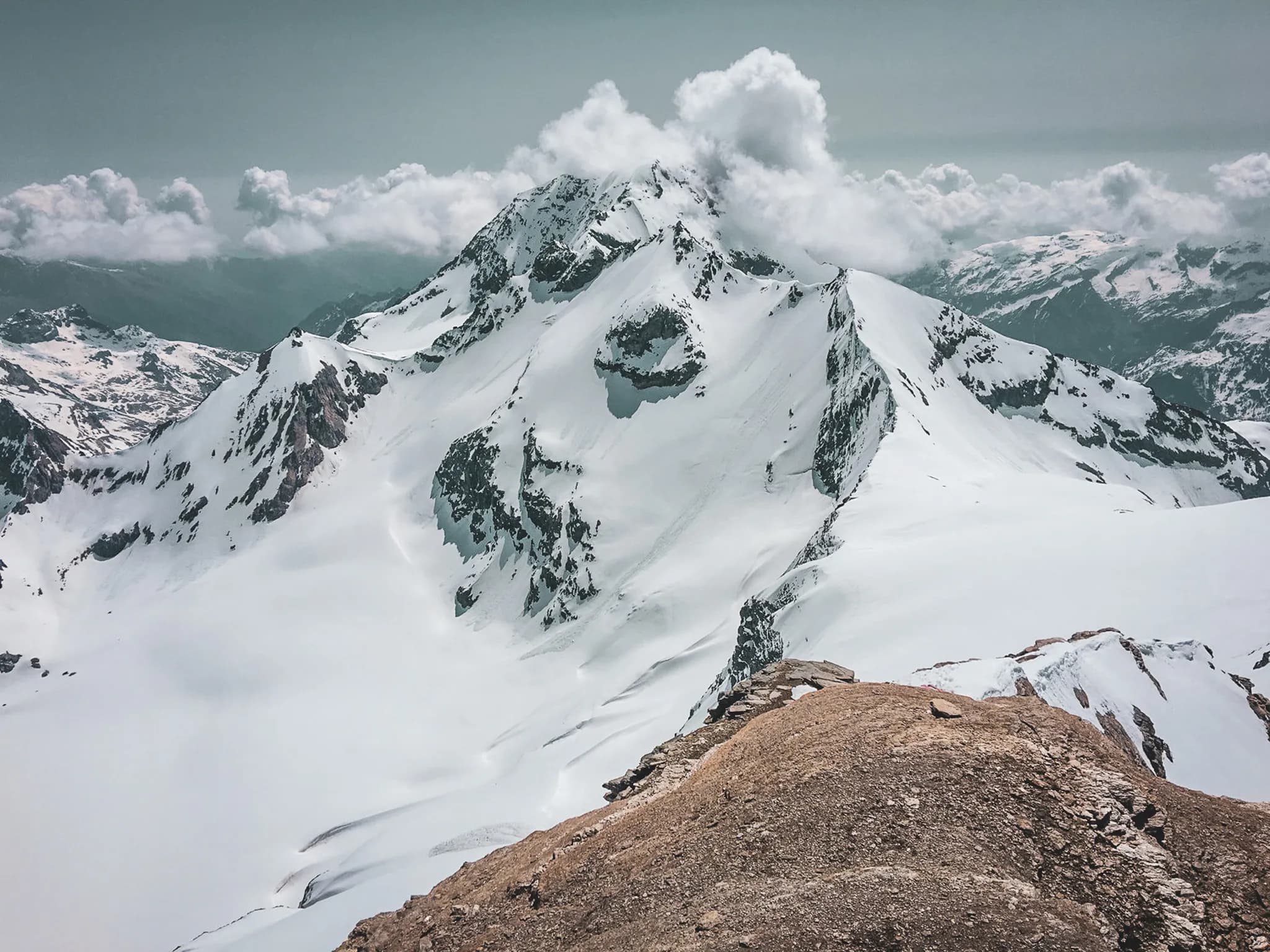 A majestic, snow-covered Alpine peak rises from an impressive mountain landscape. The slopes of the summit, dotted with rocks and snow, merge into a vast plateau.