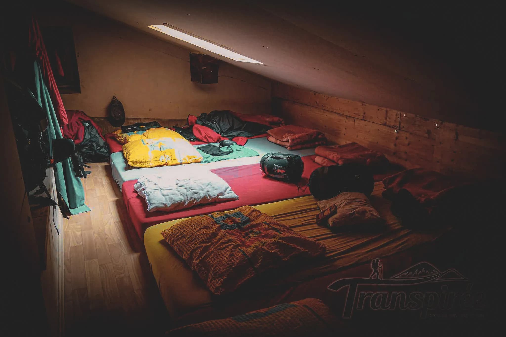 A spacious dormitory with several beds on the floor, covered with colourful mattresses and blankets. Natural light streams in through a skylight, partially illuminating the space, while the rest of the room is bathed in natural light.