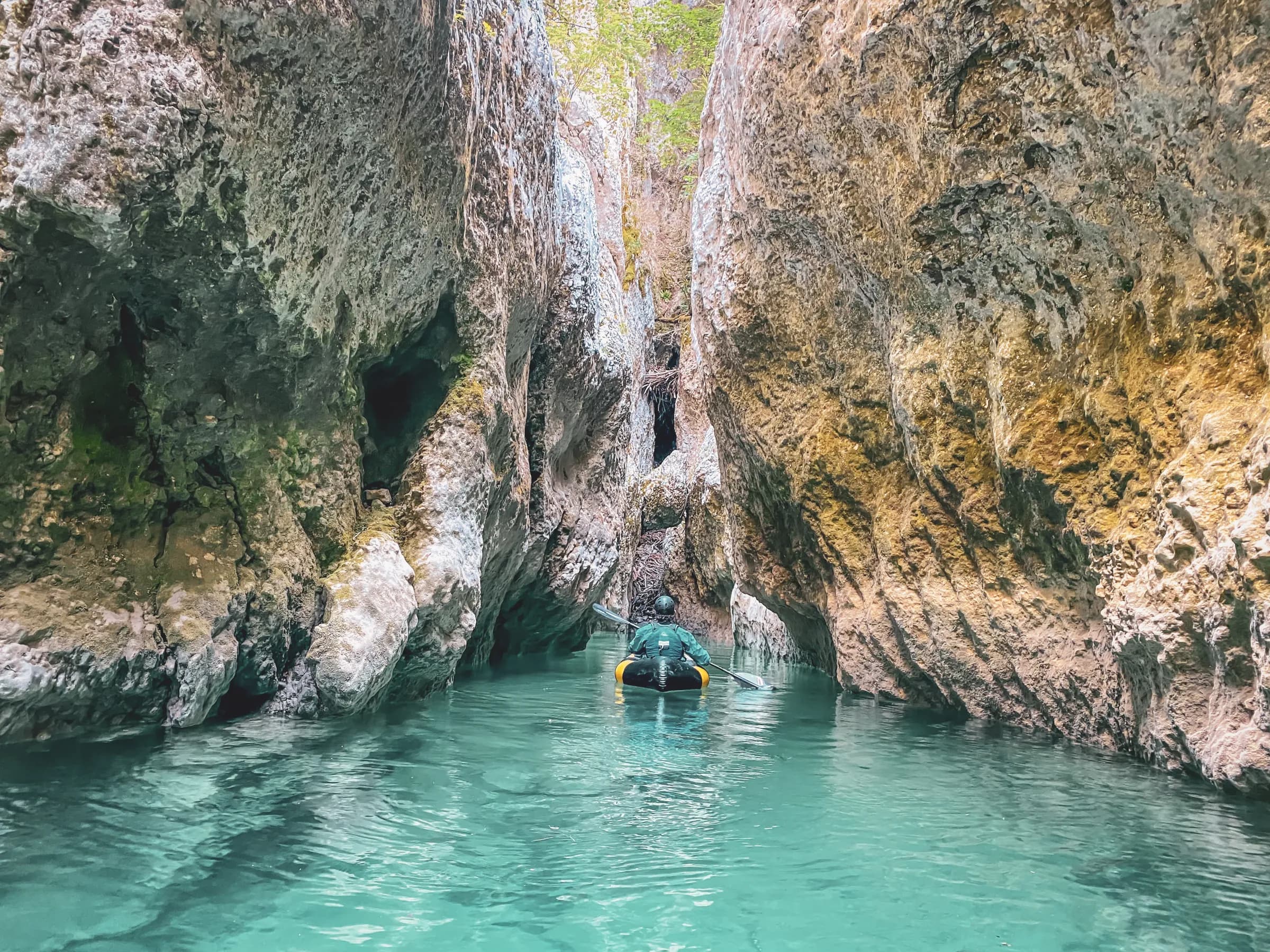 A kayaker navigates through a narrow gorge, surrounded by high rock faces of varying textures. The light green water reflects the light, creating a peaceful atmosphere. The vegetation is
